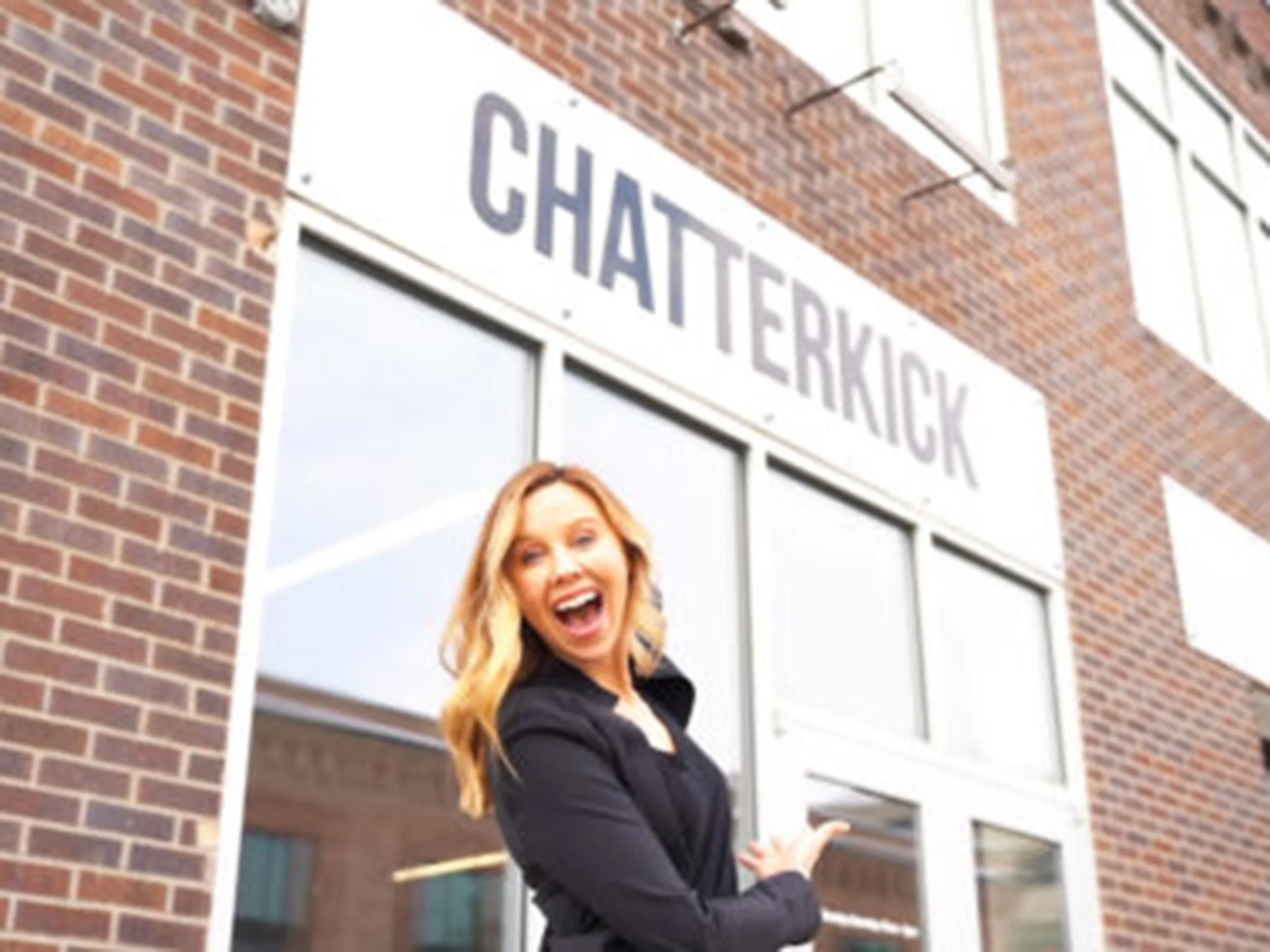 A smiling woman in a black blazer standing in front of a building with a sign that reads 'Chatterkick'