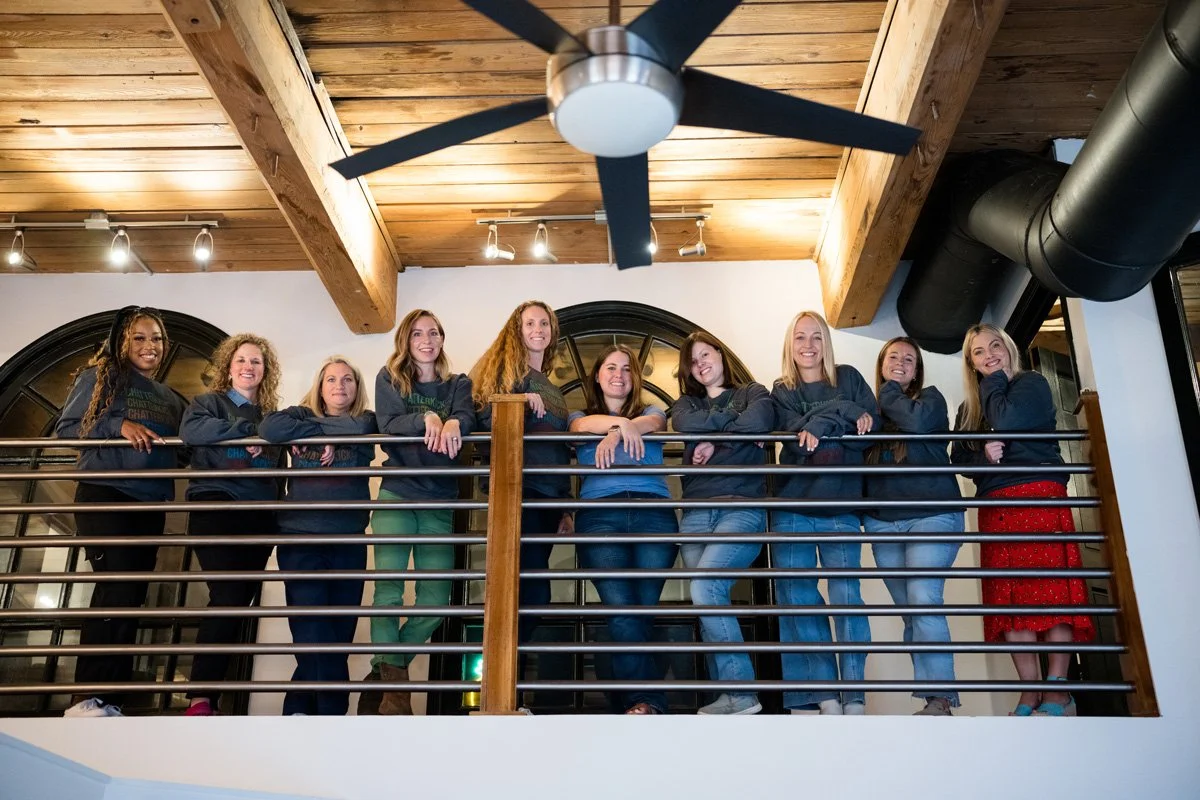 A group of nine women standing on a balcony with a wooden ceiling and black ductwork, all smiling and leaning on a metal railing.