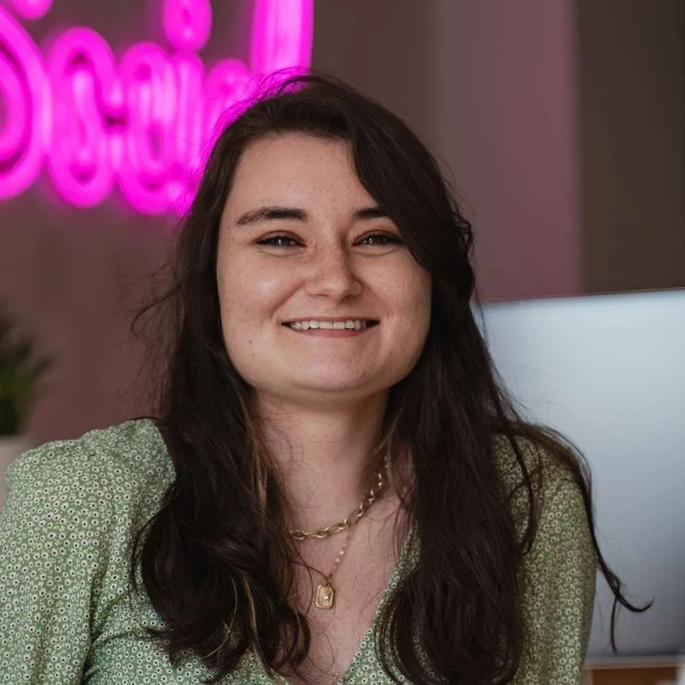 A woman with long dark brown hair smiling in an office setting with a pink neon sign in the background.