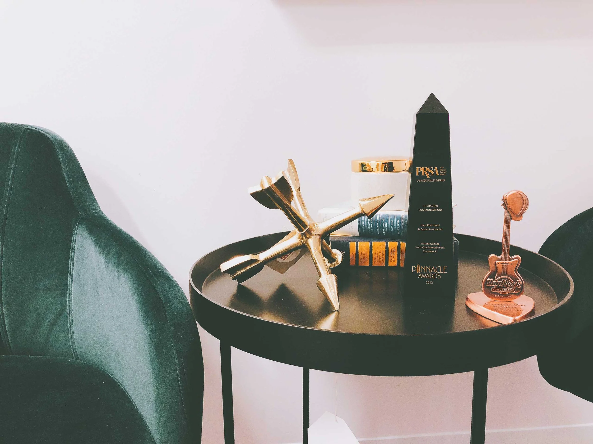 Decorative items on a black side table next to a green chair include a gold abstract sculpture, a black award trophy with gold accents, a small guitar figurine, and a stack of books, with a plain white wall in the background.