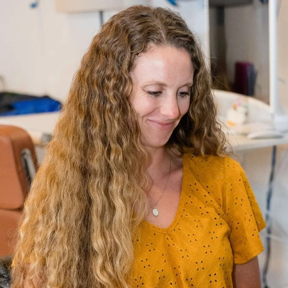 A woman with long, curly, light brown hair wearing a yellow eyelet blouse smiling softly.