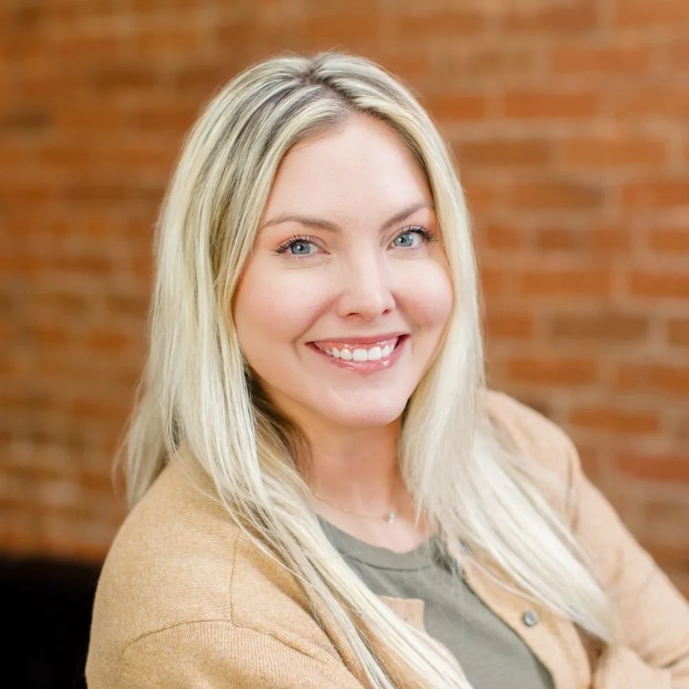 Close-up portrait of a smiling woman with long blonde hair, blue eyes, and fair skin, standing in front of a brick wall.