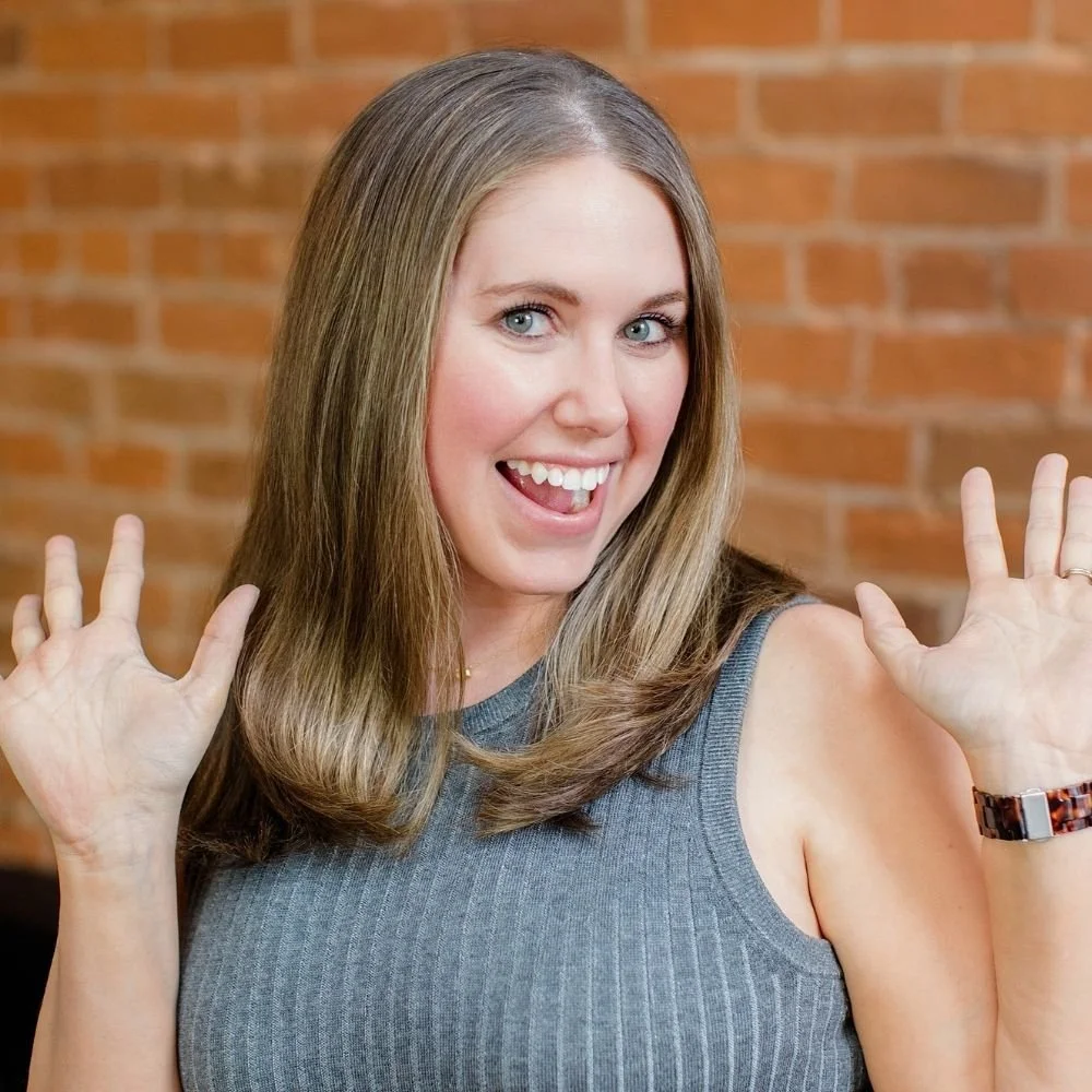 A woman with long light brown hair, blue eyes, wearing a sleeveless gray top, smiling and playfully sticking out her tongue while raising both hands in a playful gesture, standing in front of a brick wall.