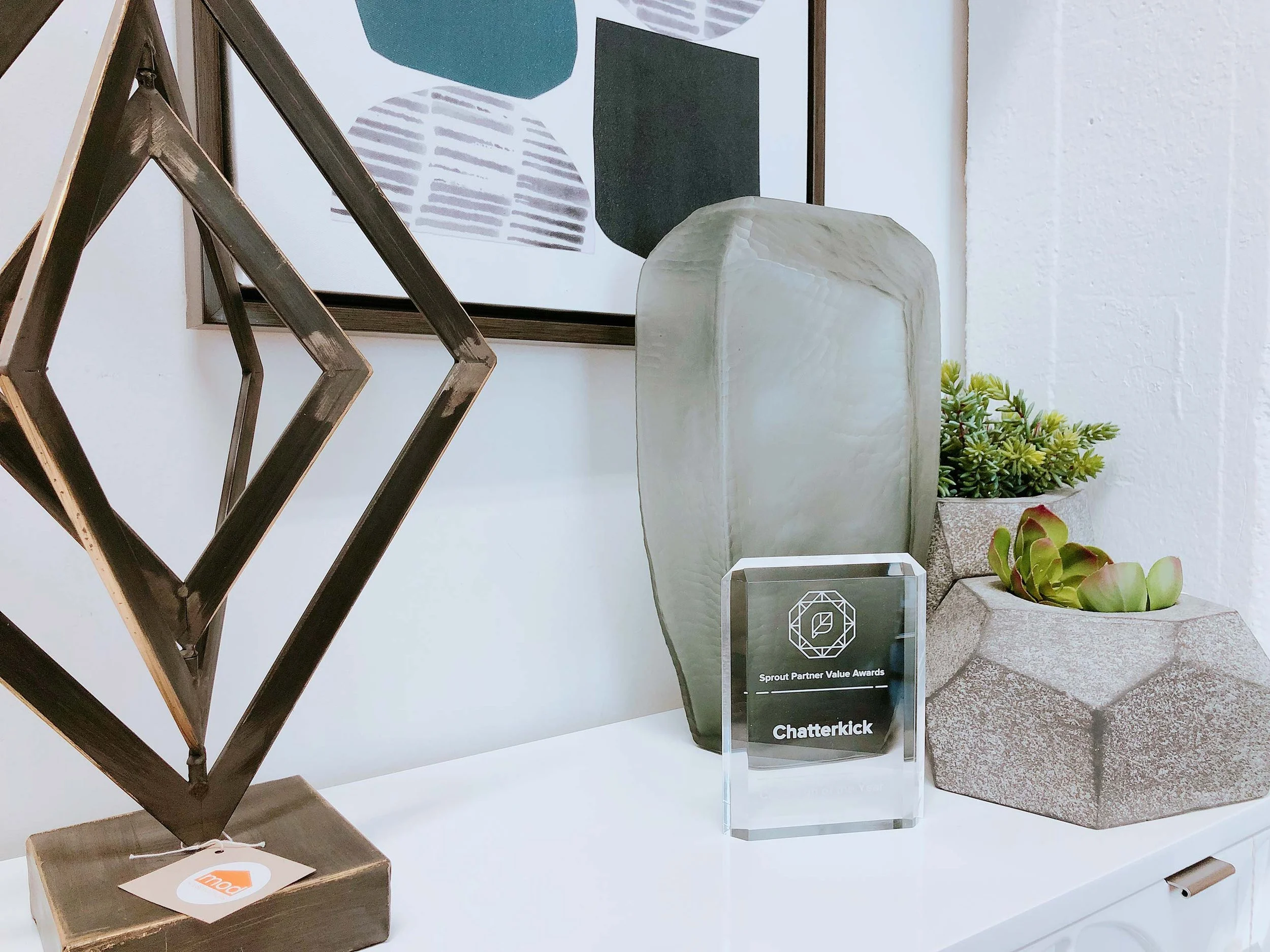 Decorative objects on a white surface including a geometric metallic sculpture, a large green vase, a framed award with the text 'Sprout Partner Value Awards Chatkerrick,' a gray stone planter with green succulents, and an abstract face art print in the background.