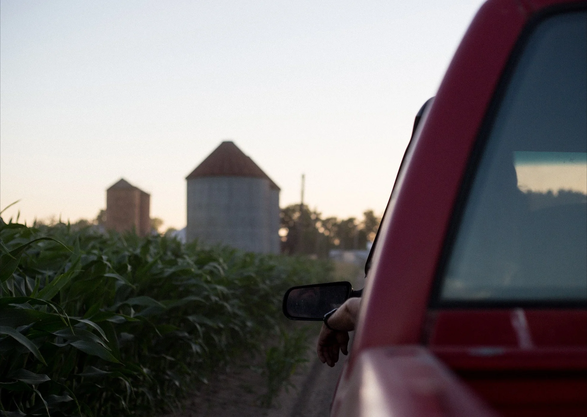 A red vehicle with a person’s arm hanging out the window, driving past green crops with farm silos in the background at sunset.