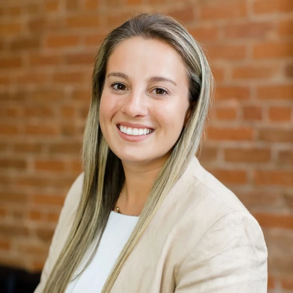 A woman with long blonde hair, smiling, wearing a light-colored blazer and white top, standing in front of a brick wall.