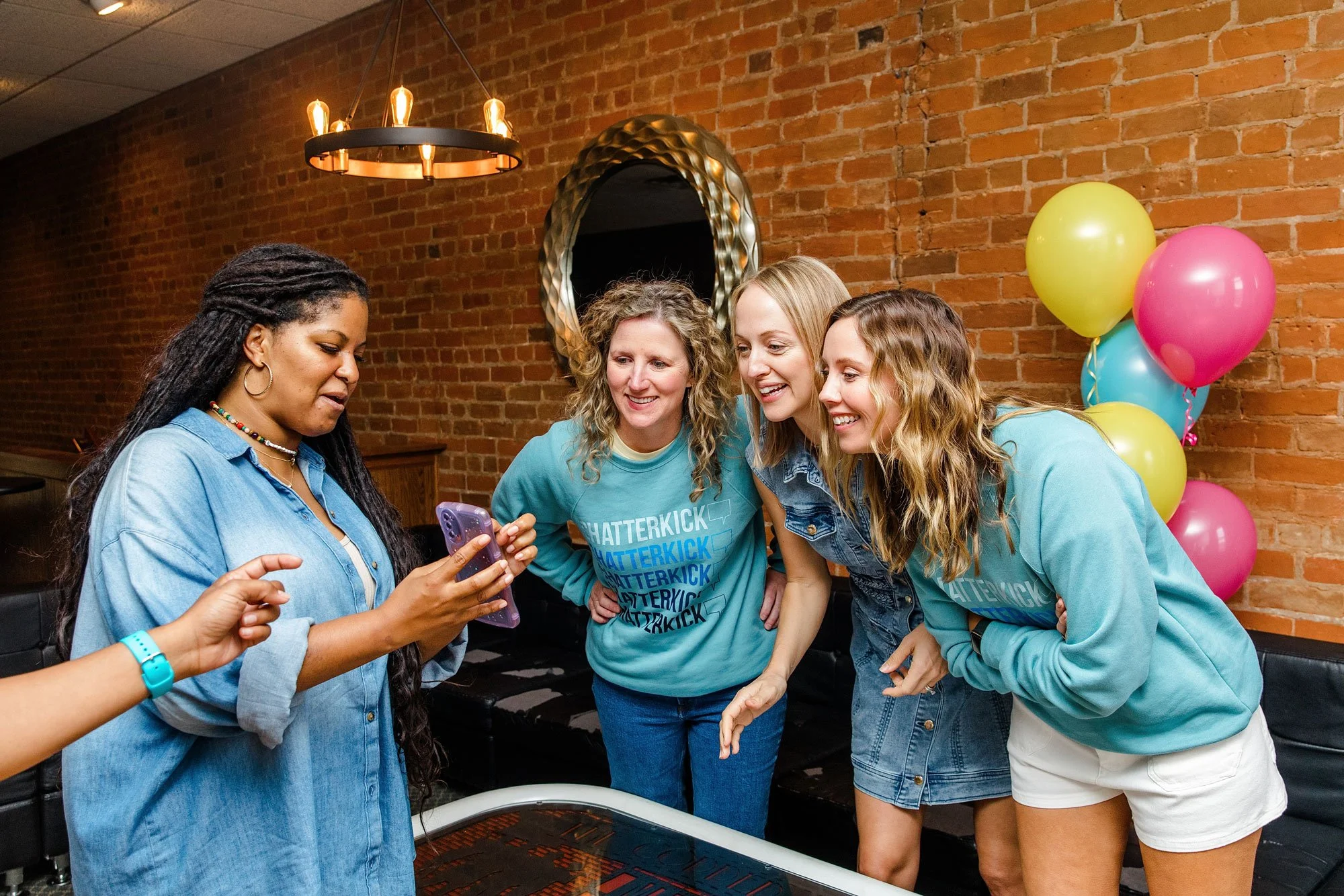 Four women celebrating a birthday at a party with pink, yellow, and blue balloons, looking at a phone and smiling.
