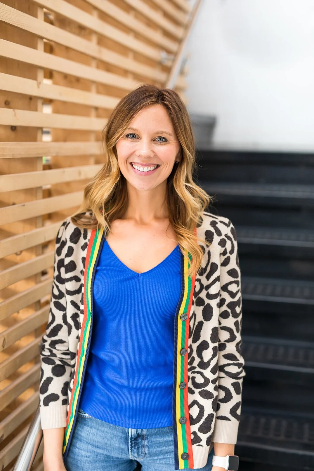 A woman with blonde wavy hair smiling, wearing a blue top, leopard print cardigan with rainbow-colored trim, and jeans, standing indoors near a wooden slat wall and black stairs.