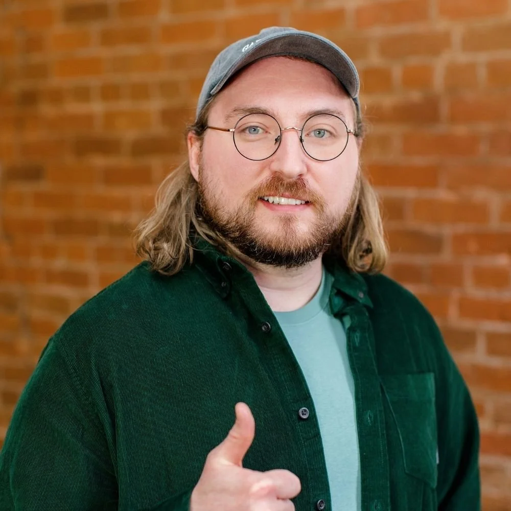 A man with long hair, glasses, and a beard wearing a gray cap, black shirt, and green jacket, smiling and giving a thumbs-up in front of a brick wall.