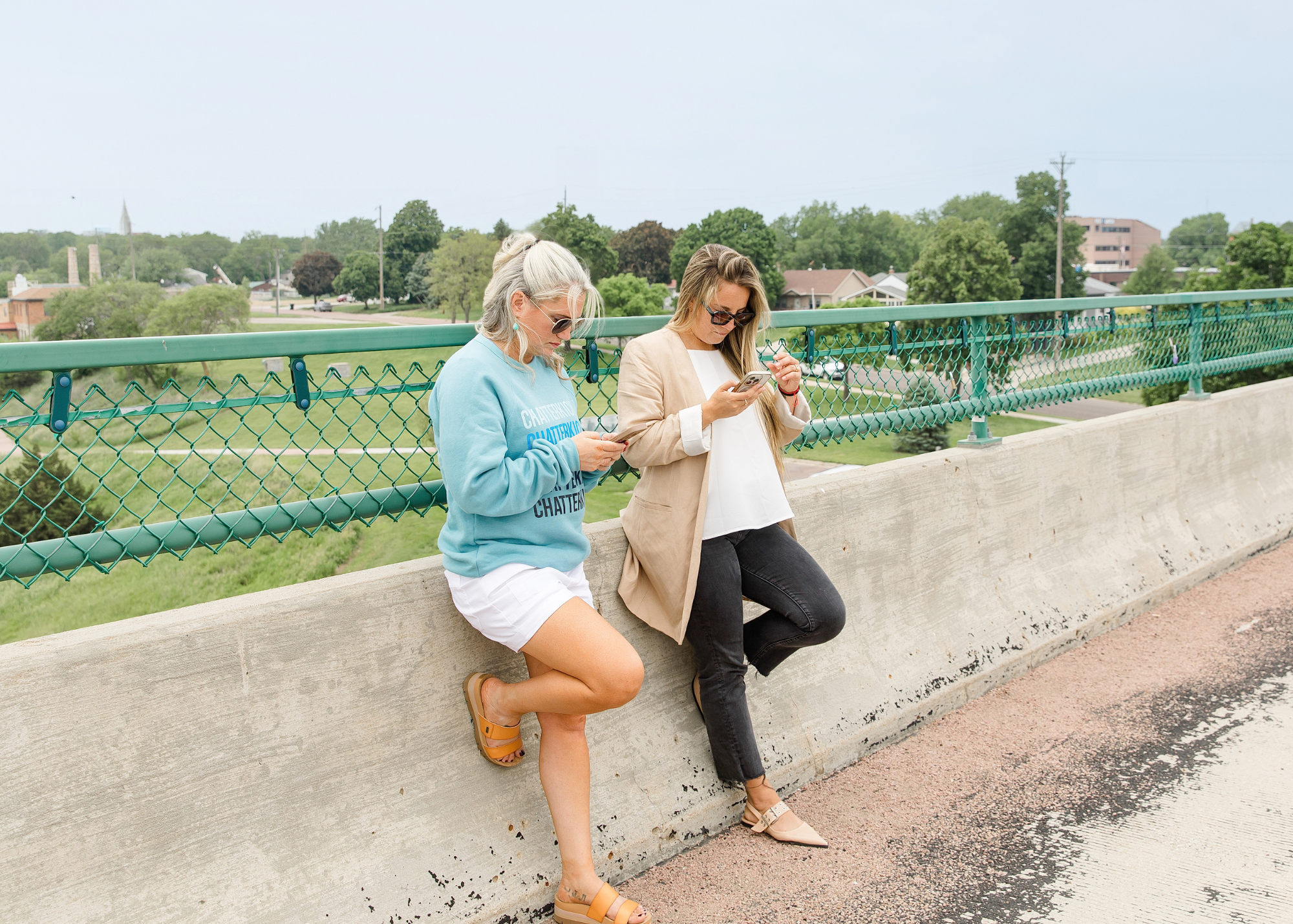 Two women leaning against a concrete railing while looking at their phones on a bridge with a green chain-link fence, overlooking a green park and buildings in the distance on a cloudy day.