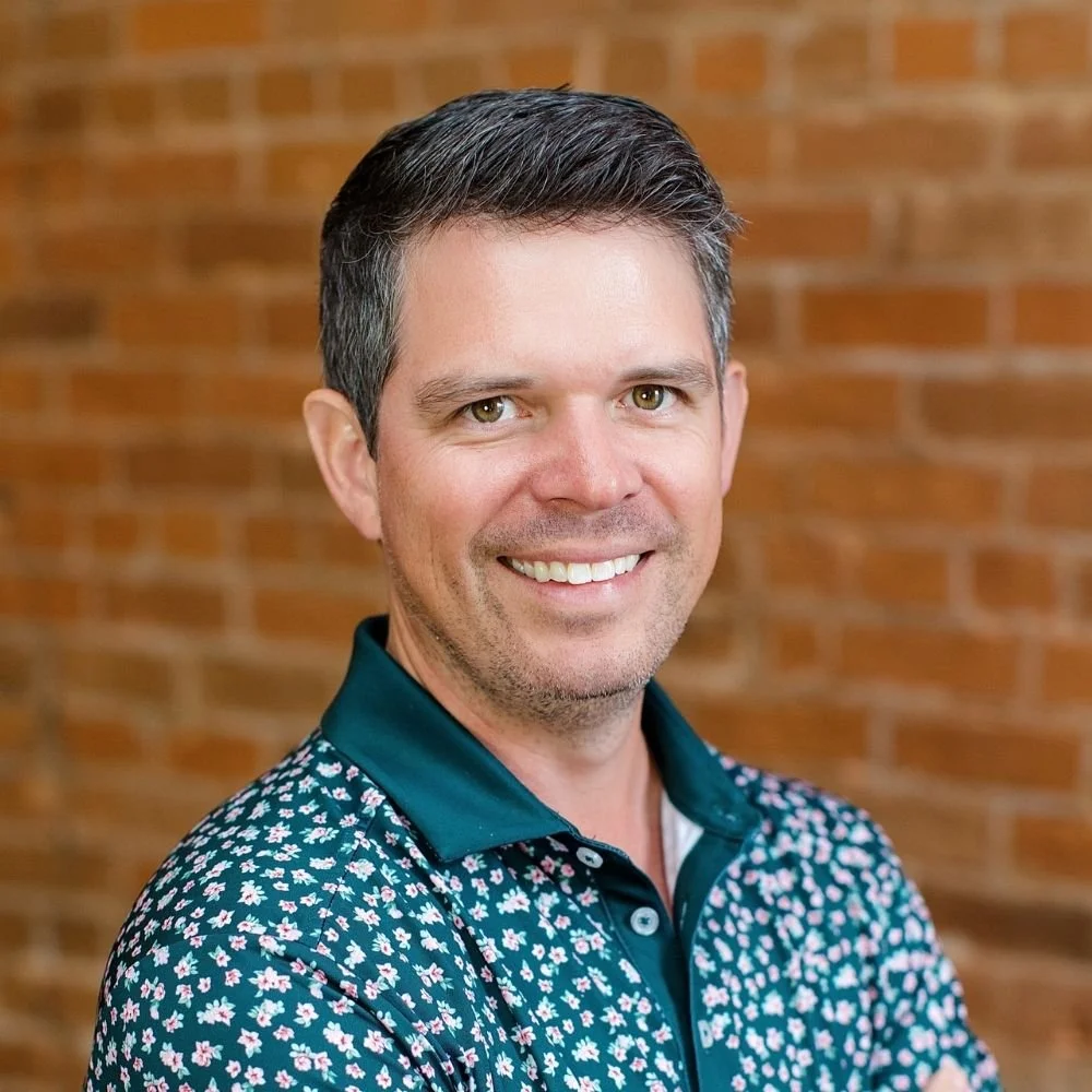 A smiling man with short dark hair, wearing a floral patterned shirt, stands in front of a brick wall.