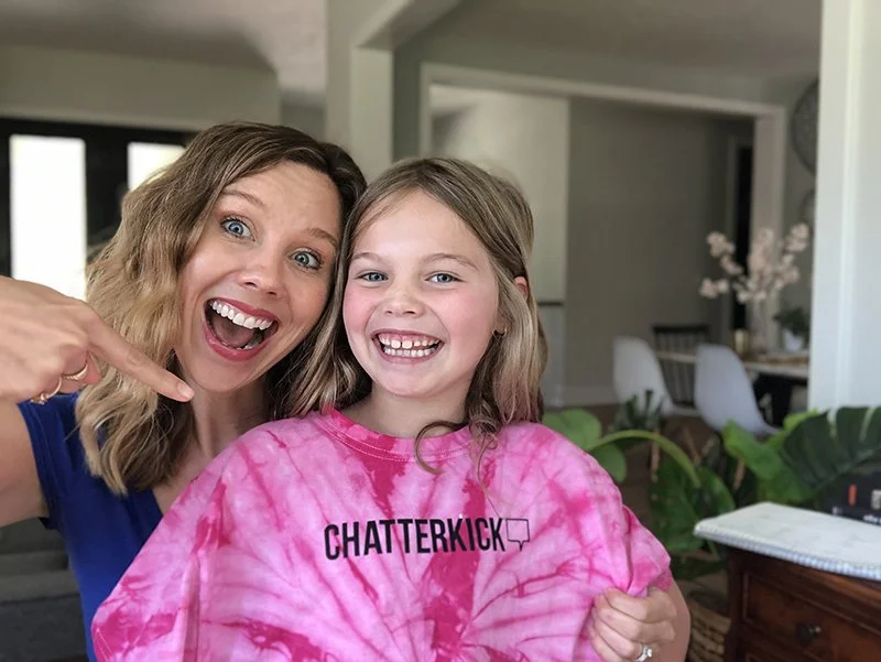A woman and a girl smiling happily indoors, with the woman pointing to the girl, both wearing casual clothes.