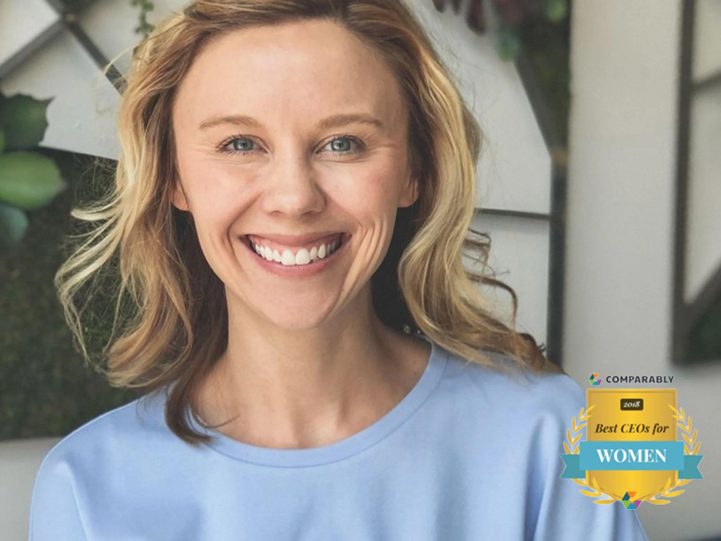Smiling woman with curly blonde hair wearing a light blue shirt, standing outdoors with greenery and a white wall in the background. There is a badge on the bottom right that says '2018 Best CEOs for Women' from Comparably.