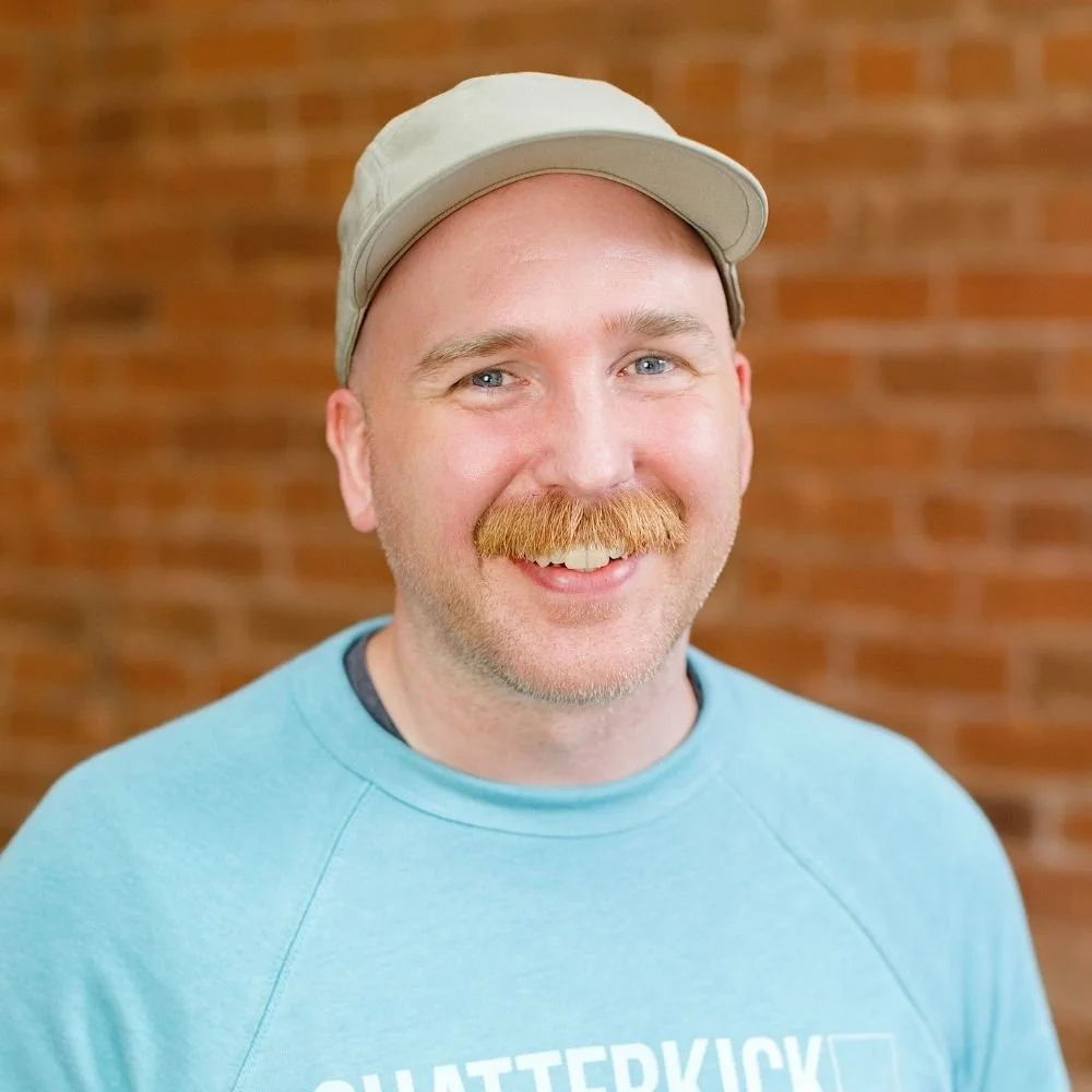 A smiling man with a beard and mustache wearing a light gray cap and a blue sweatshirt in front of a brick wall.