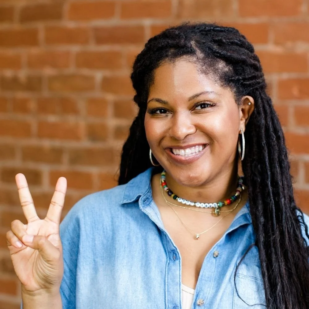 Smiling woman with dreadlocks making a peace sign, wearing a denim shirt, earrings, and necklaces with a brick wall background.