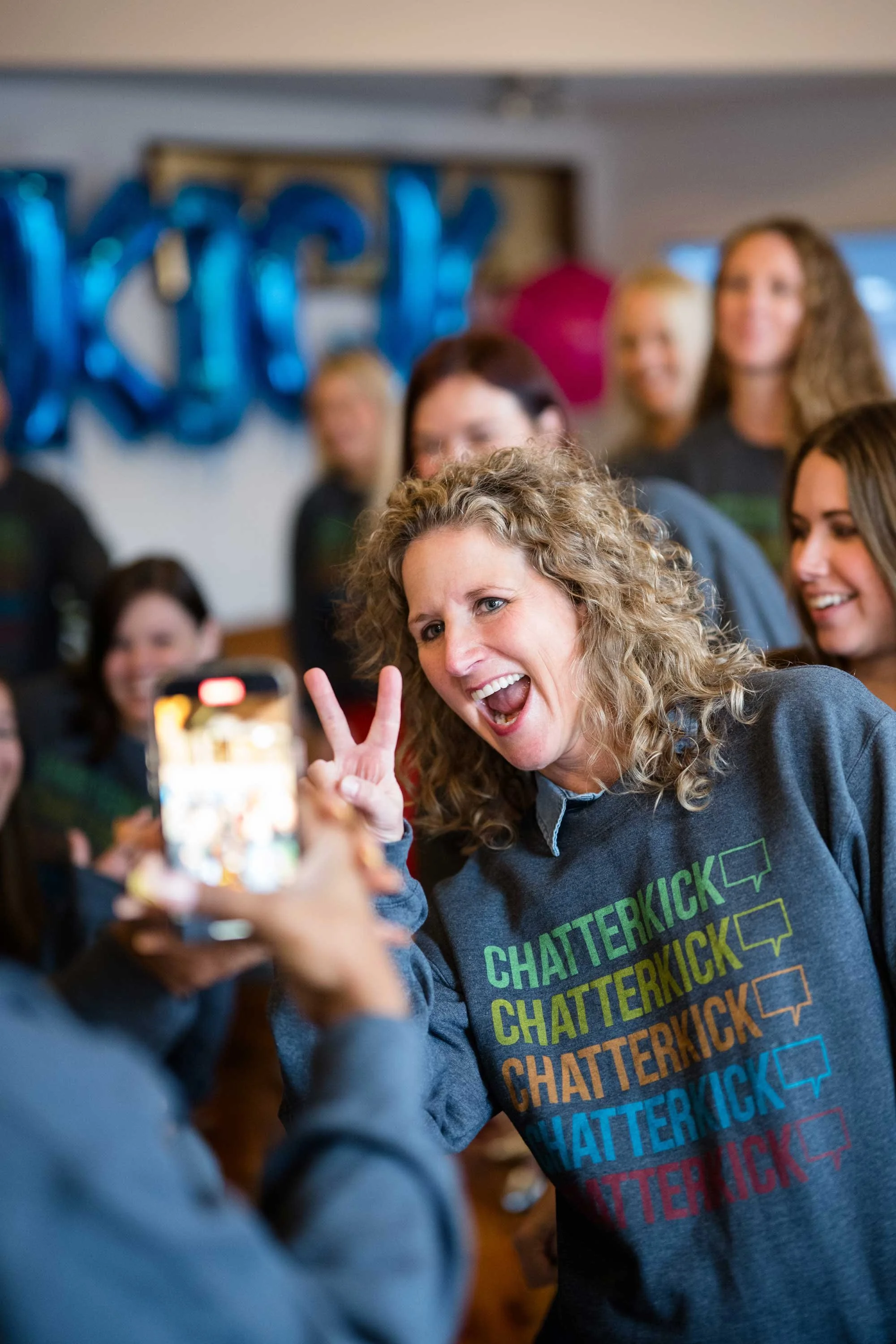A woman with curly blonde hair, wearing a gray sweatshirt with colorful text 'CHATTERKICK,' is posing with a peace sign while someone takes her photo.