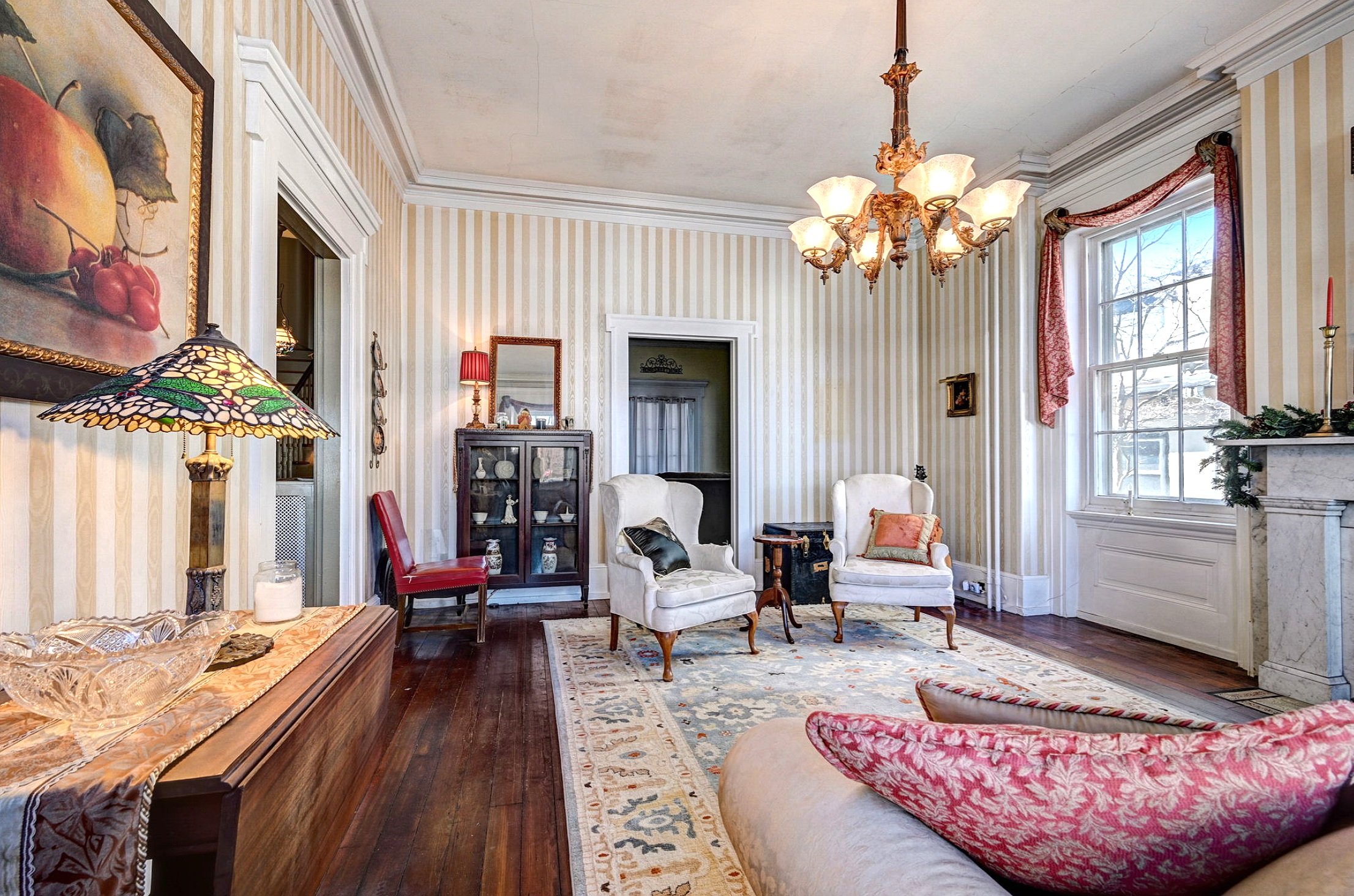 Interior of a vintage living room with striped wallpaper, antique furniture, a chandelier, and decorative items on display.