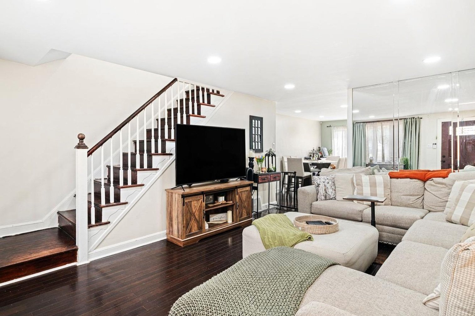 Living room with beige sectional sofa, green throw, and wooden TV stand. Staircase with dark wood steps and white railing. Dining area with table and chairs in the background. Hardwood floors and large windows with teal curtains.