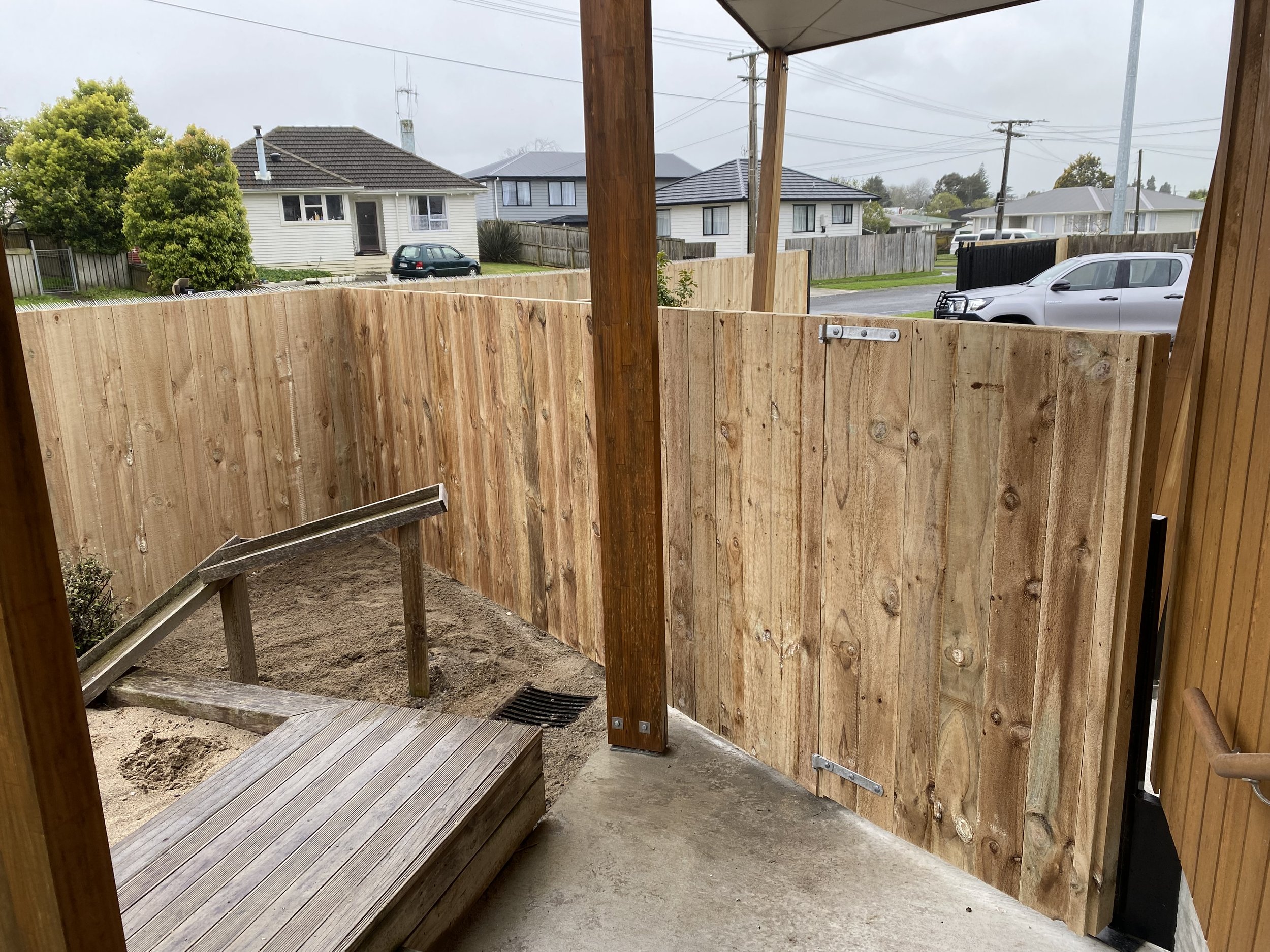 View from a covered porch with wooden railings overlooking a backyard. There is a new wooden fence enclosing the area, and a small ramp leading to a sandy ground. Residential houses and a street with parked cars are visible in the background.