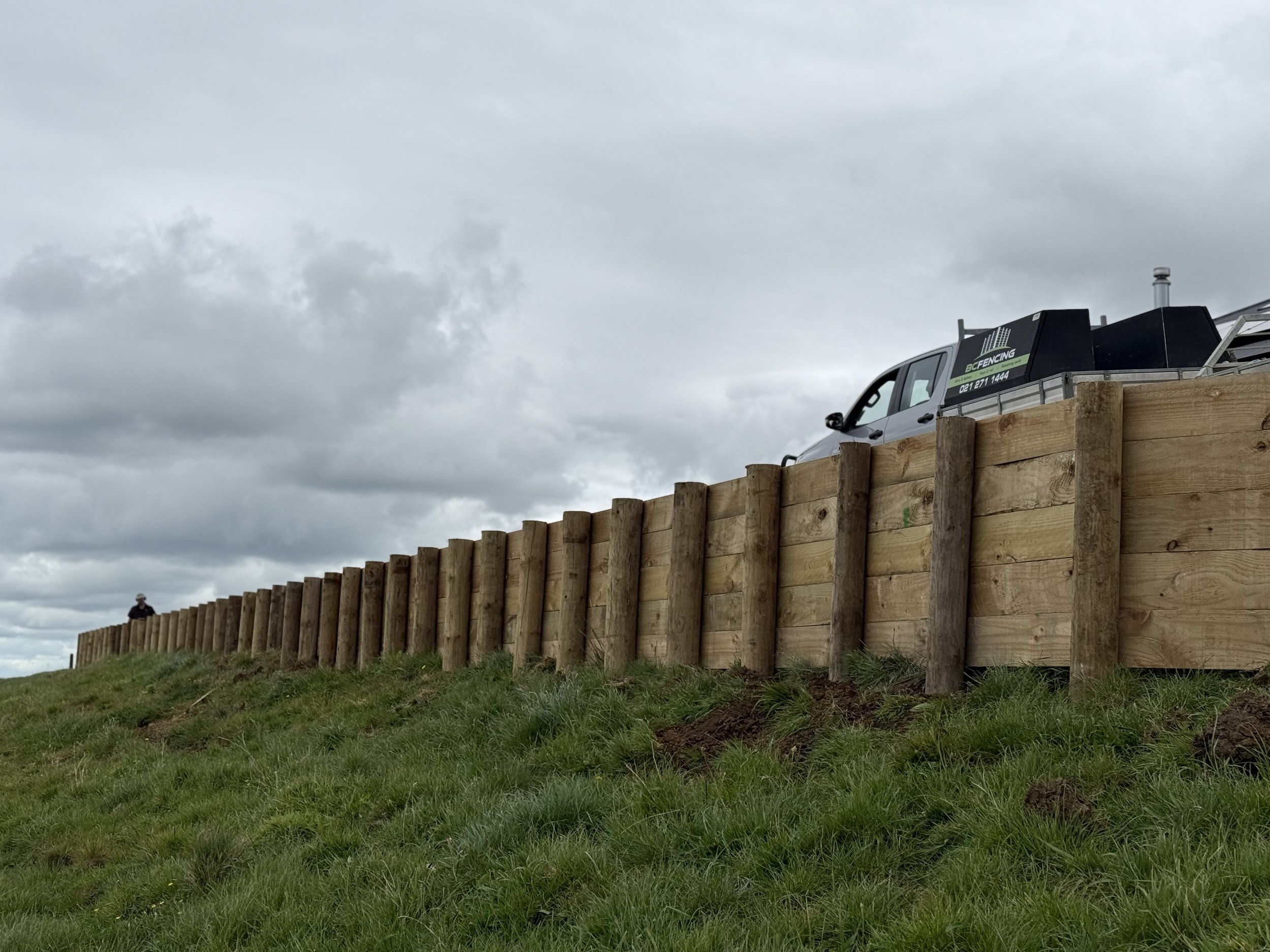 A solid retaining wall crafted from wood stands on a grassy hill with a cloudy sky in the background. A silver truck is partially visible, parked behind the fence.