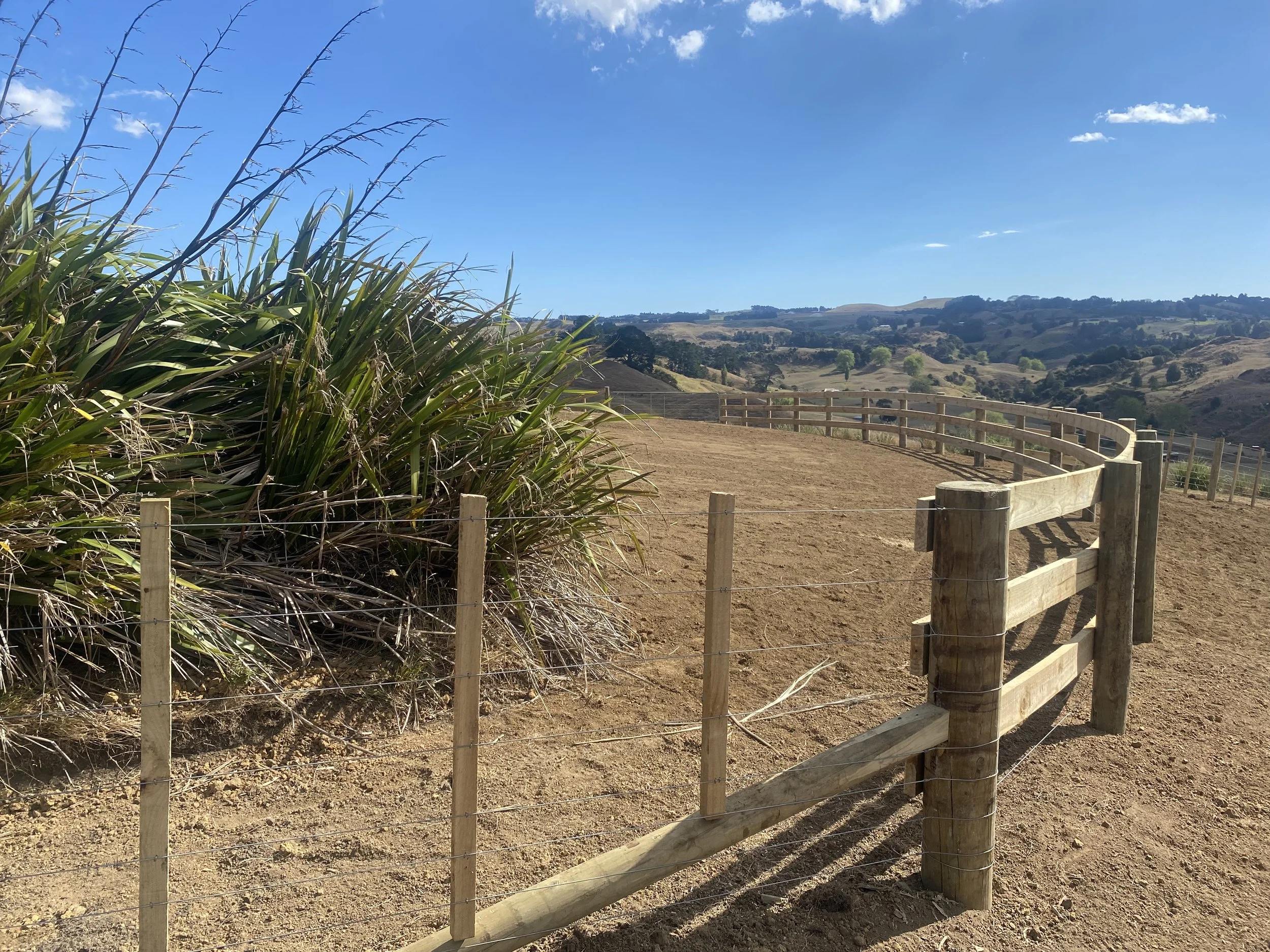 Wooden fence on a dirt hill with plants and distant hills under a clear blue sky.