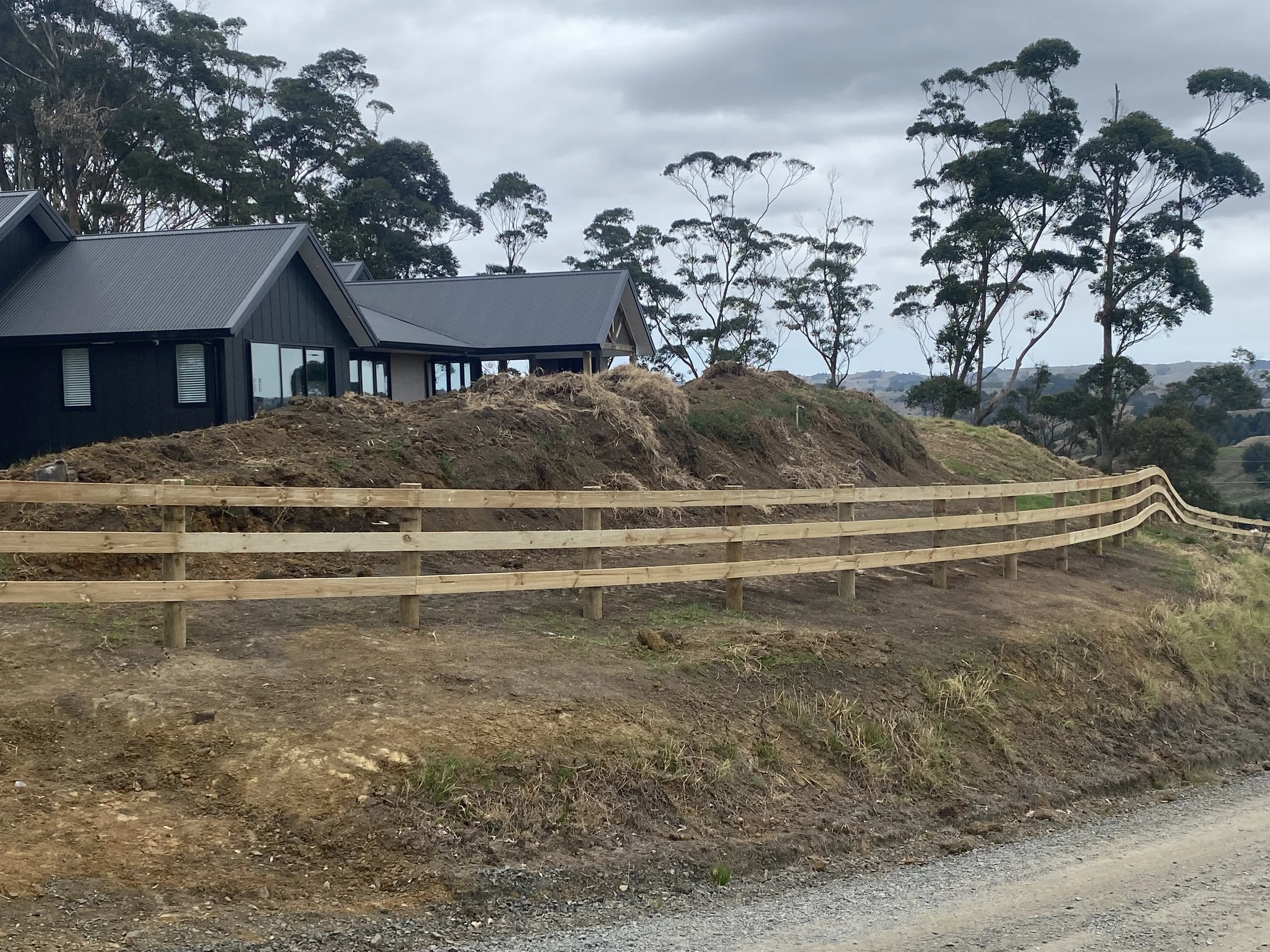 Modern house on a lifestyle block on a hill with a curved wooden post and rail fence and surrounded by trees.