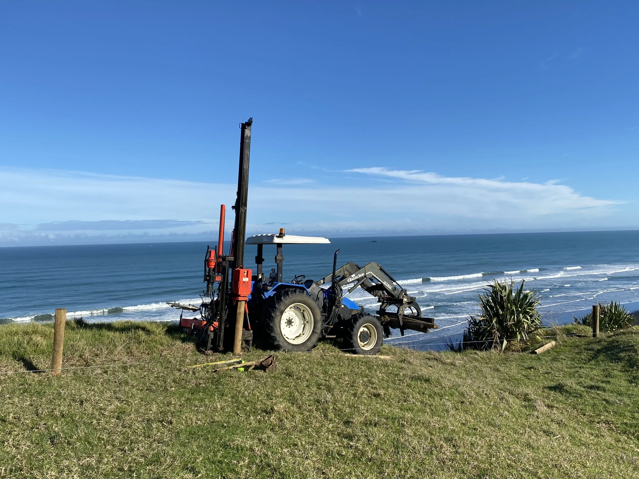 Tractor with fencing equipment on grassy cliff overlooking ocean waves under blue sky.