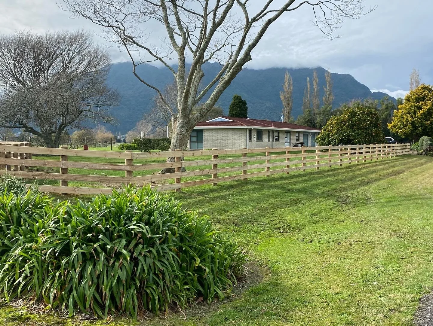 Countryside landscape with a house, wooden fence, trees, and mountains in the background.