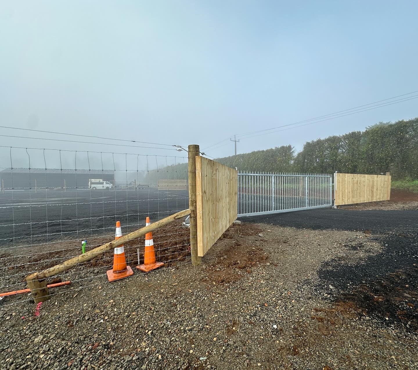 Construction site with a gravel road, wooden fence, metal gate, and orange traffic cones in foggy weather.