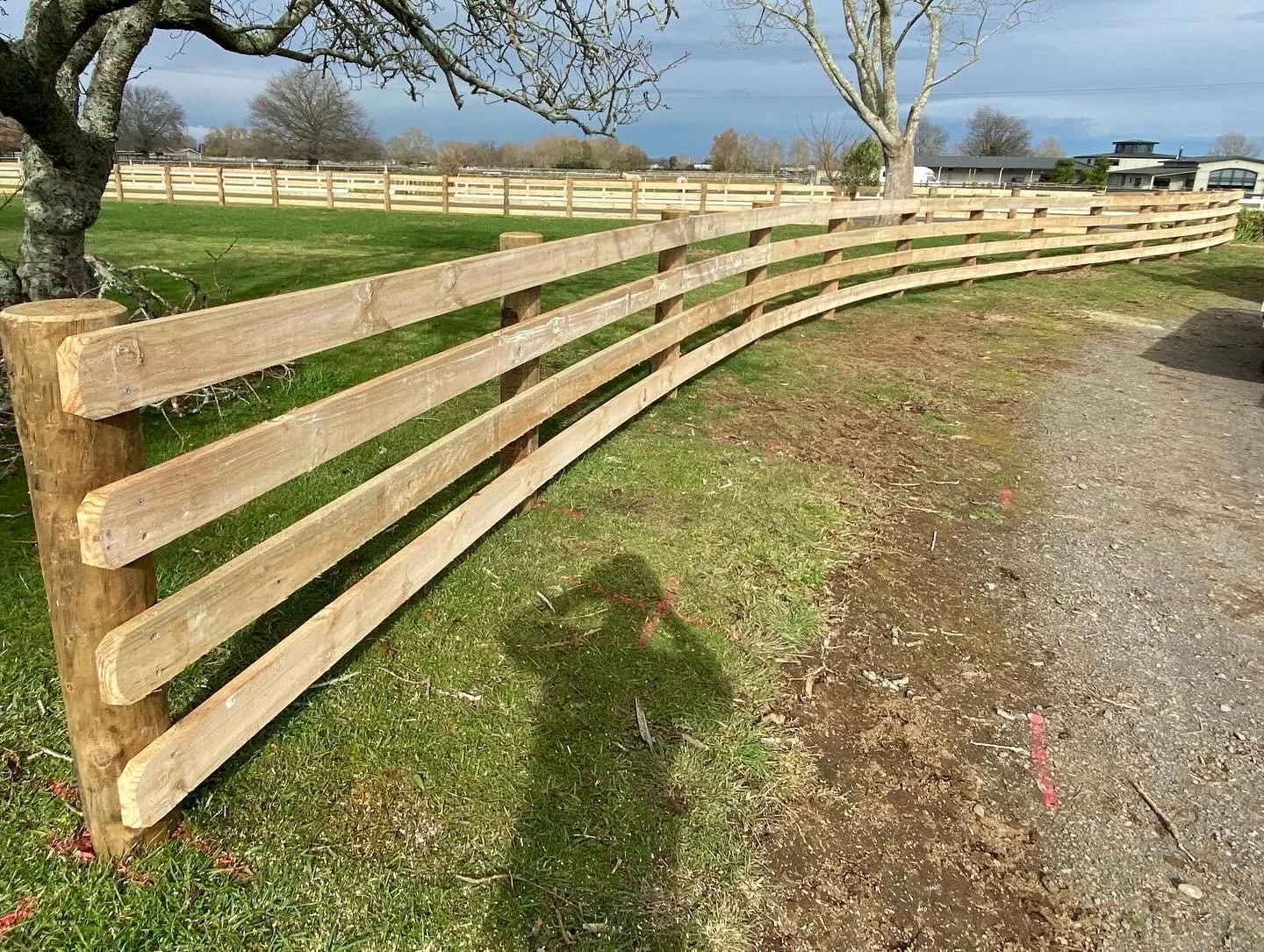 Wooden post and rail fence on grass, with trees and a cloudy sky in the background.