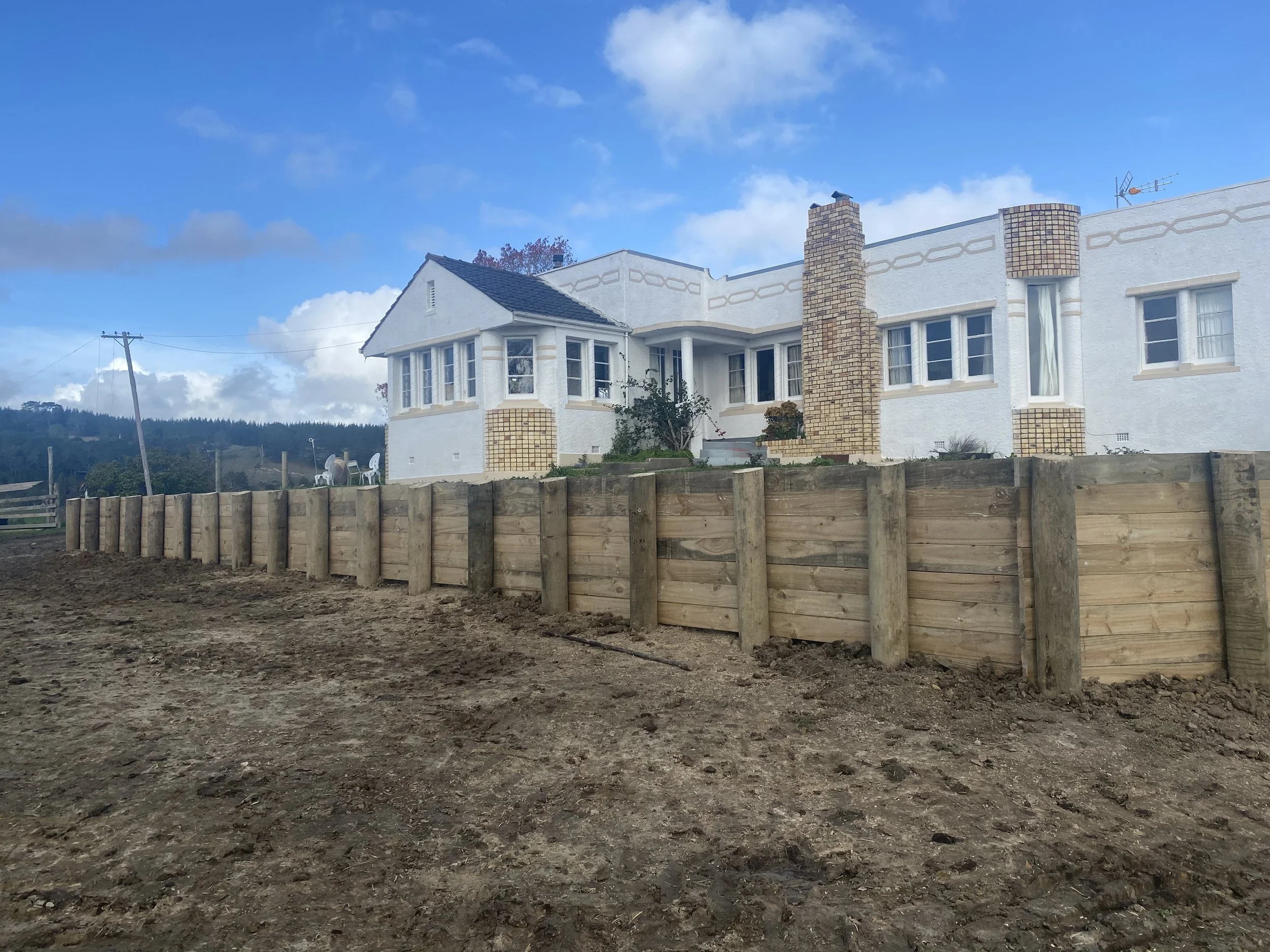 White stucco house on a hill with wooden retaining wall, chimney, and cloudy sky.