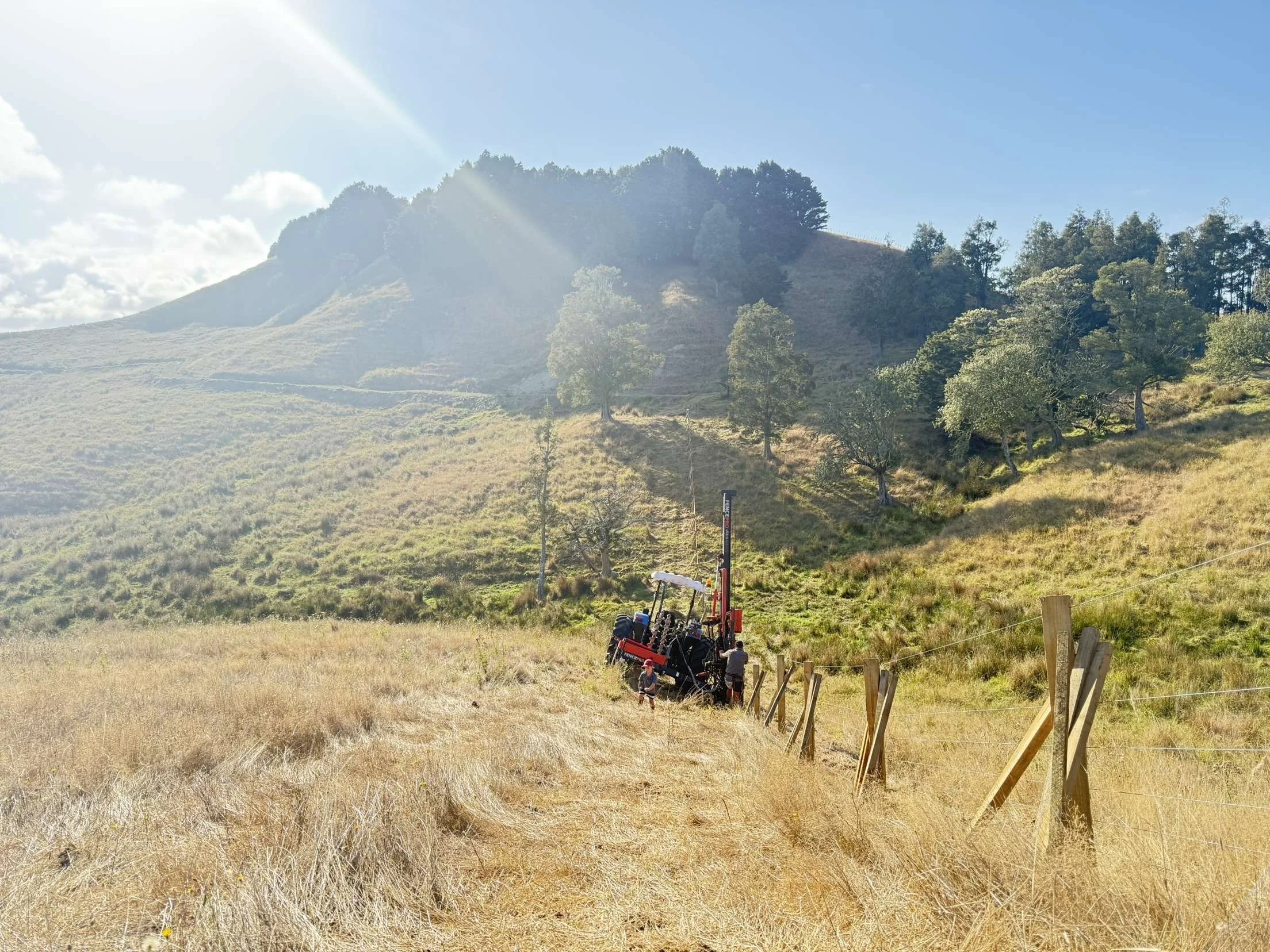A tractor in a sunny rural landscape with hills, grass, and trees in the background. A wooden fence runs along the field in a differing terrain.