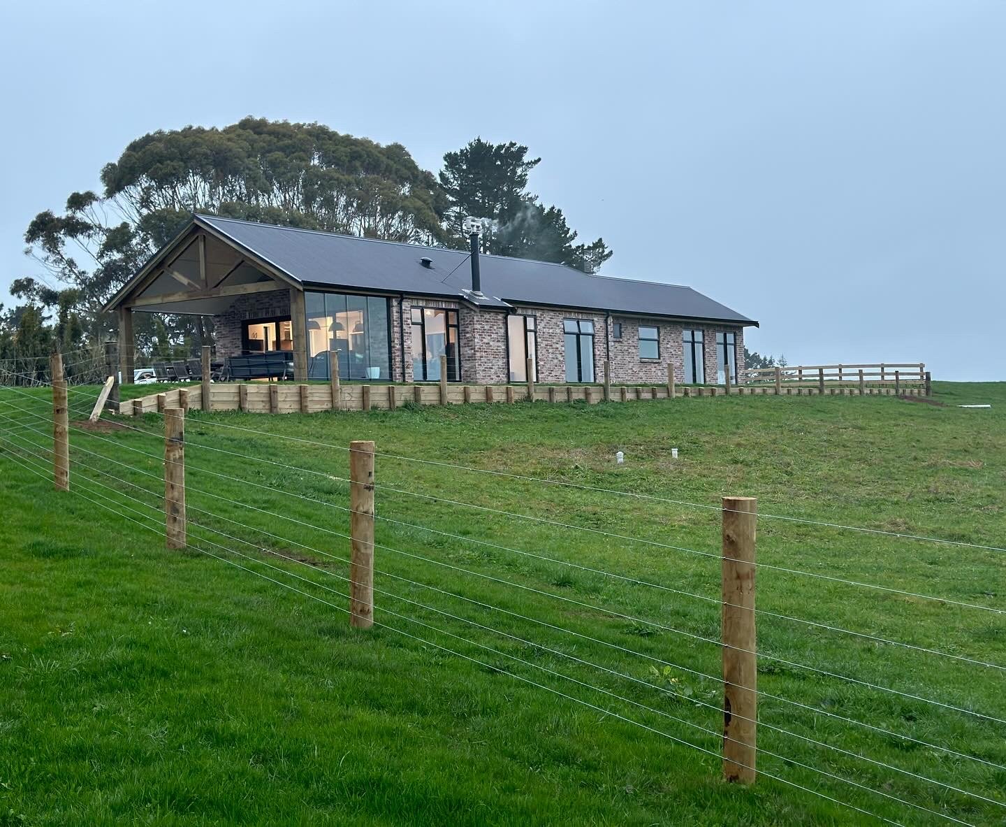 Modern farmhouse on a grassy hill with large windows built retaining wall and a fenced yard.