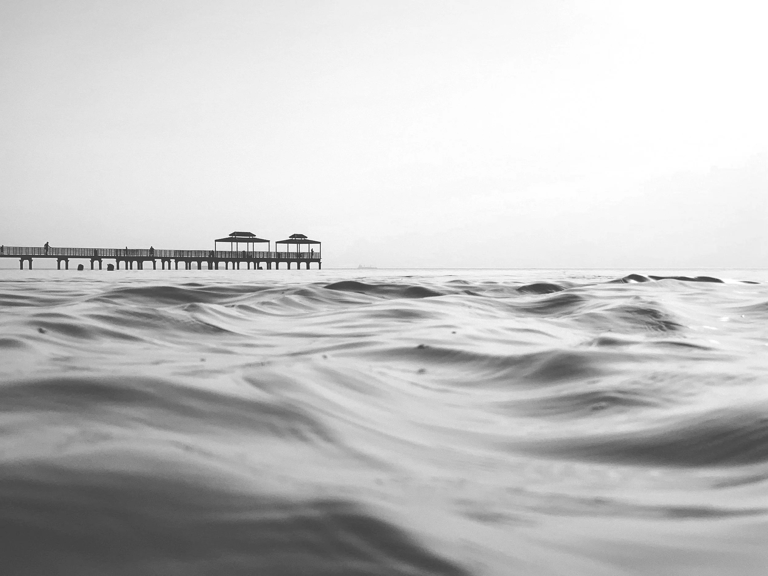 Black and white photo of a pier extending over the ocean with gentle waves in the foreground and a clear sky in the background.