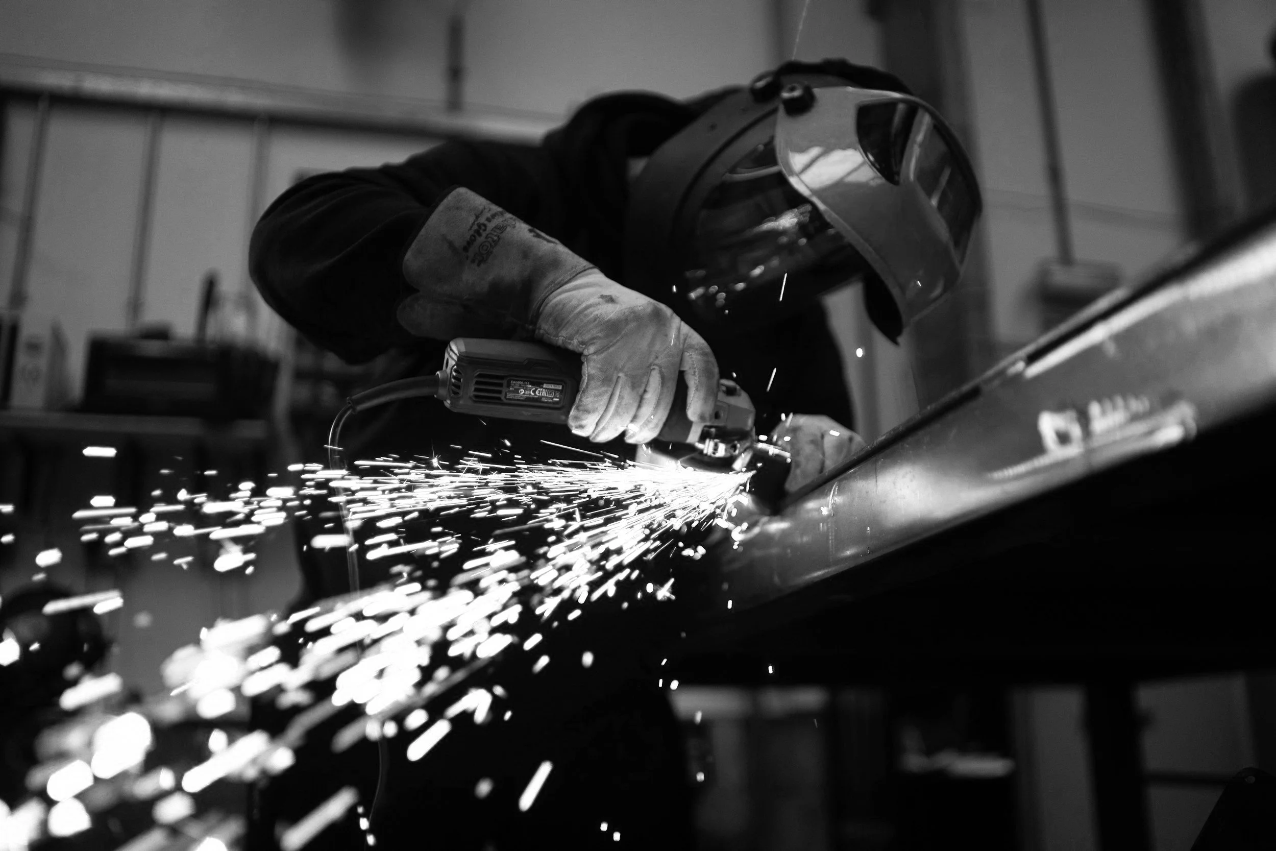 Black and white photo of a person using a grinder in a workshop, wearing protective gloves and a face shield, with sparks flying.