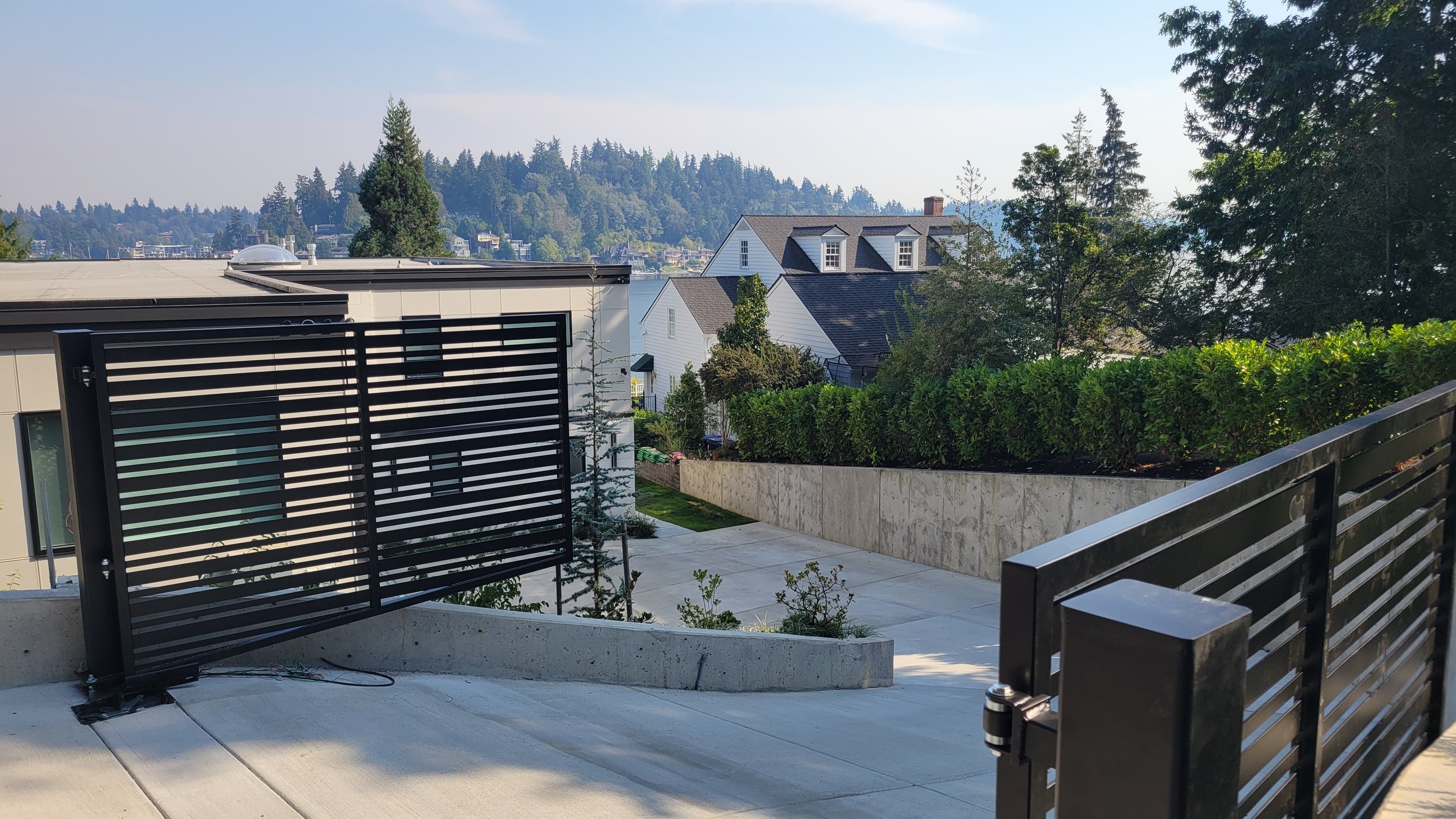 Modern fence with horizontal slats next to a paved driveway, trees in background, suburban houses visible, clear sky.