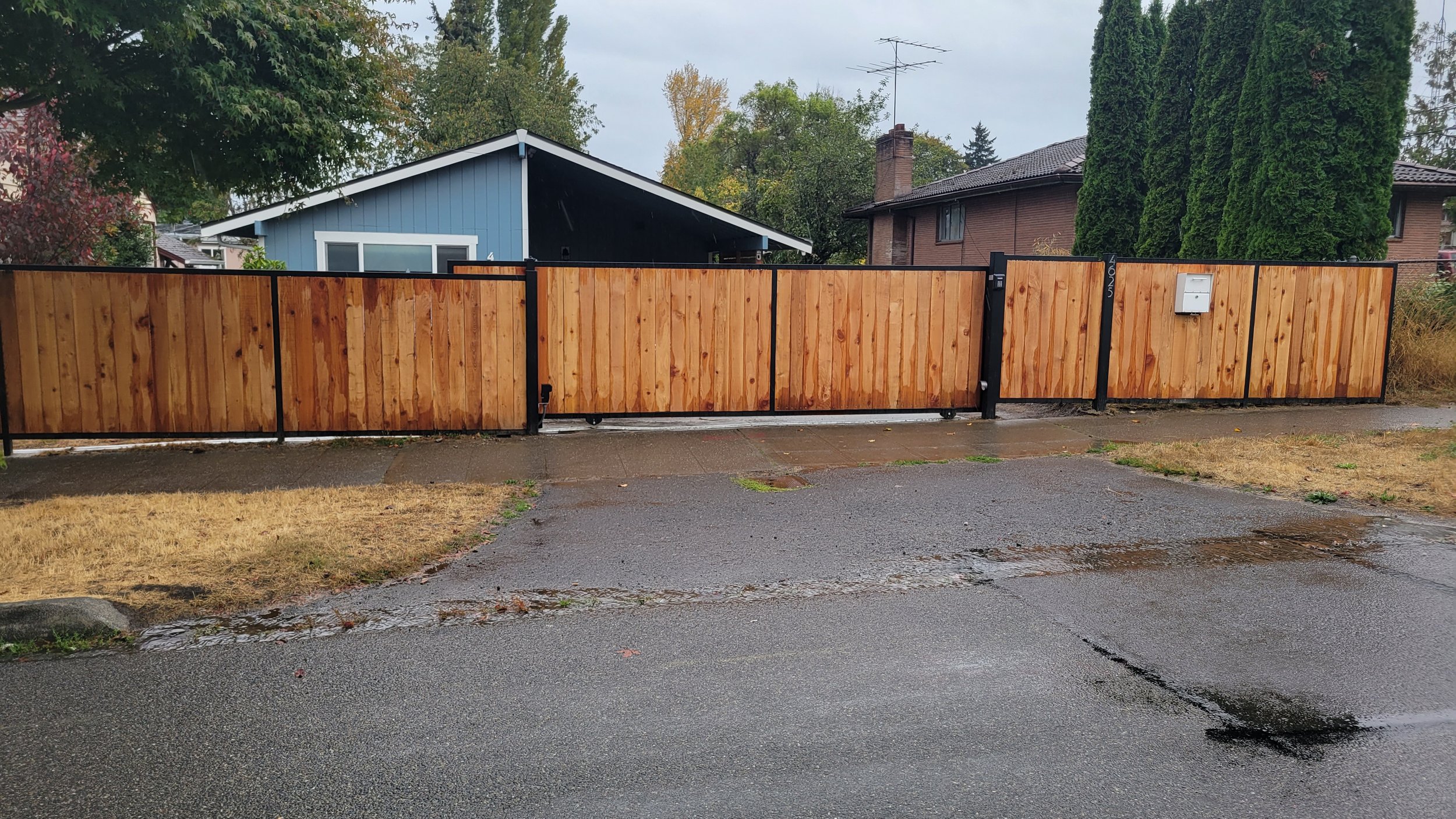 Wooden gate and fence in front of a blue house with a driveway, surrounded by green trees and overcast sky.