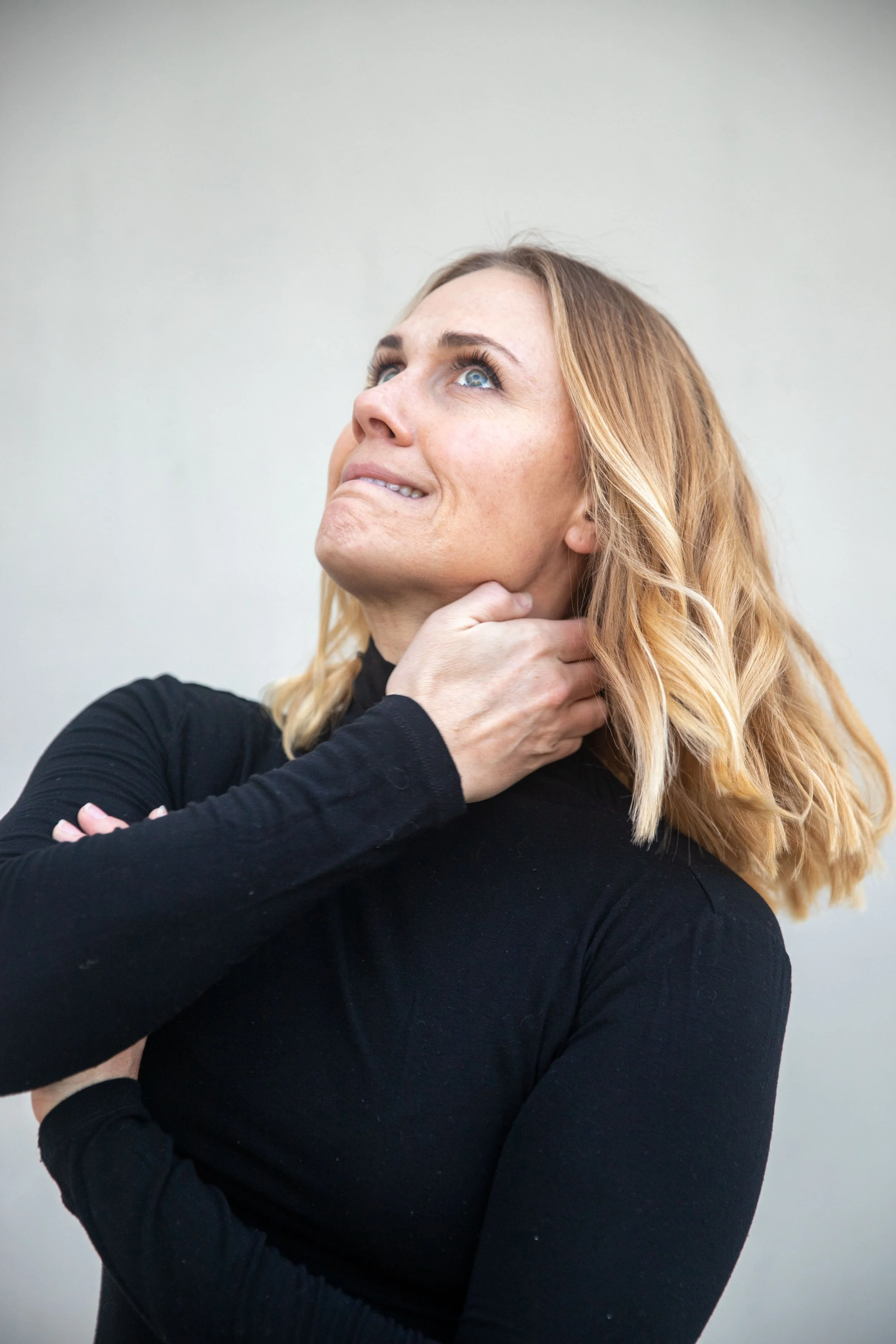 Alison Shurter wearing a black long sleeve shirt, holding her neck and looking upward, against a plain light-colored background.