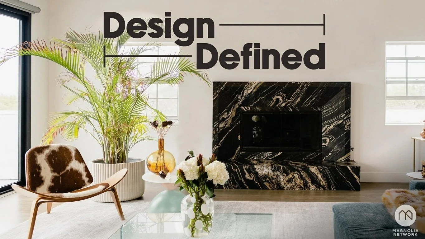 Modern living room with large potted plant, cowhide chair, glass vase with white flowers, black marble fireplace, and large windows with white walls, with the text "Design Defined" in bold black letters on the wall.