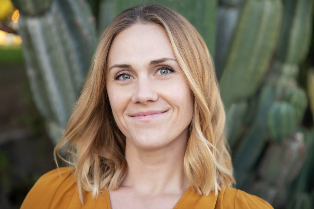 A woman with shoulder-length blonde hair, blue eyes, and a slight smile standing outdoors near large green cactus plants.