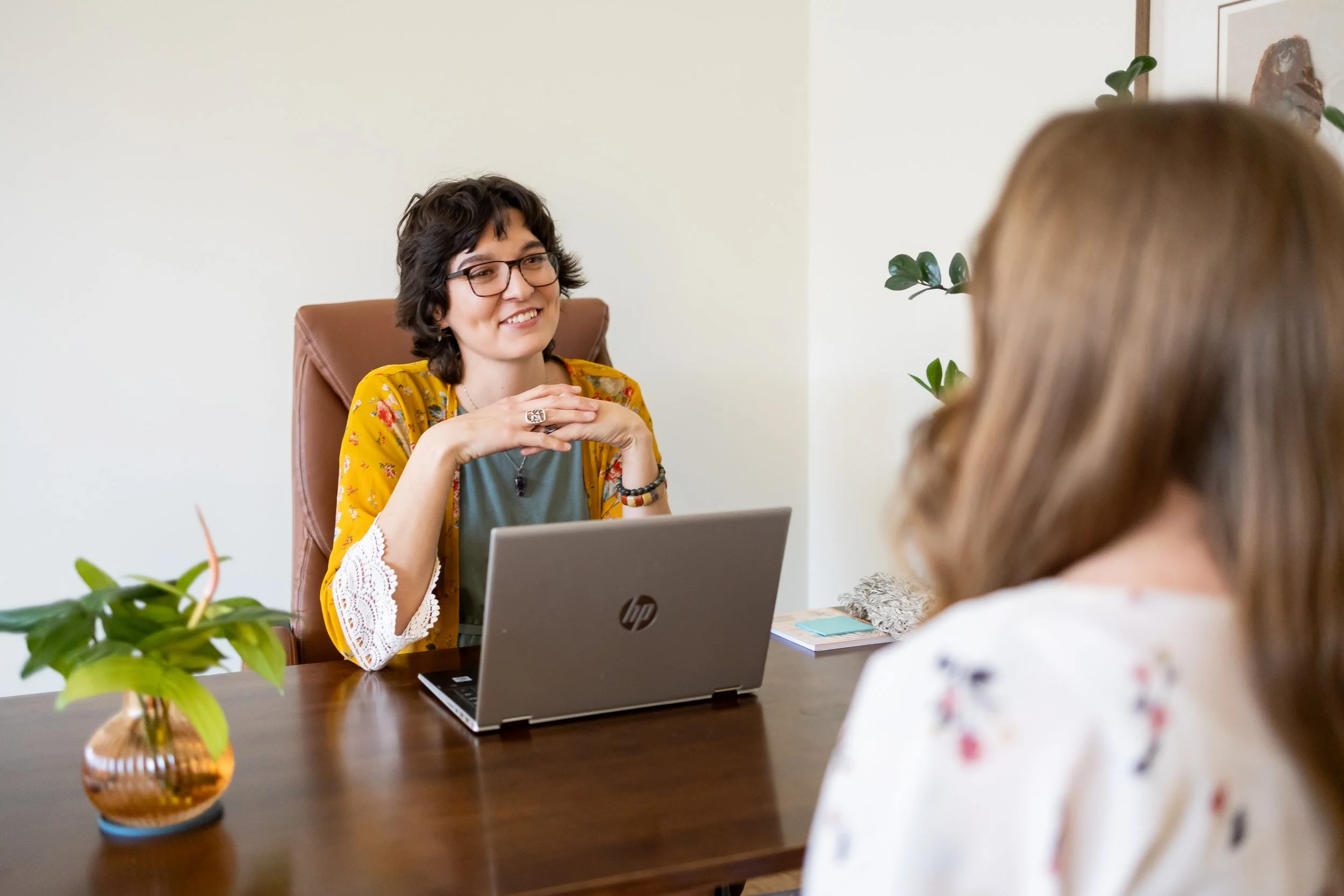 Dr. Bobi Farrow, licensed acupuncturist and naturopathic doctor, sitting at her desk in Lincoln office, speaking with a client (back of head visible), with open laptop, notebook, sticky notes, and a bundle of white sage on the desk