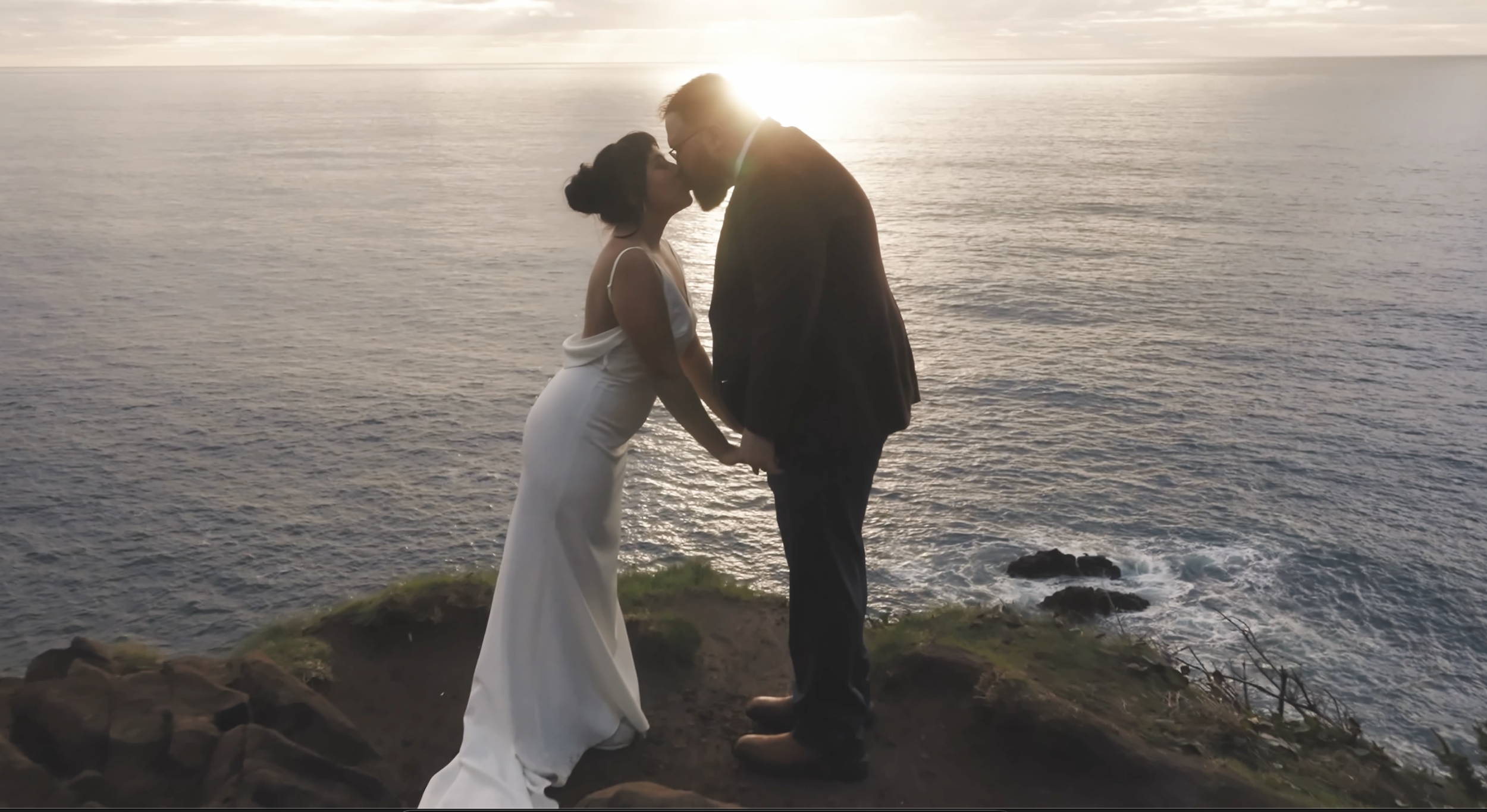 A couple in wedding attire kissing by the ocean at sunset.