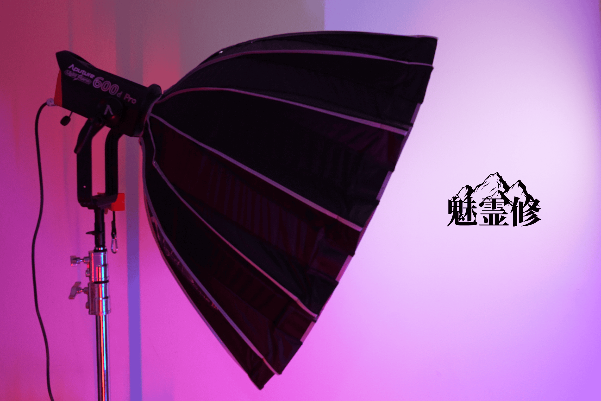 A photography lighting setup with a large umbrella reflector and a studio light mounted on a stand, illuminated with purple and pink lighting. There are Chinese characters and mountain illustrations in the bottom right corner.