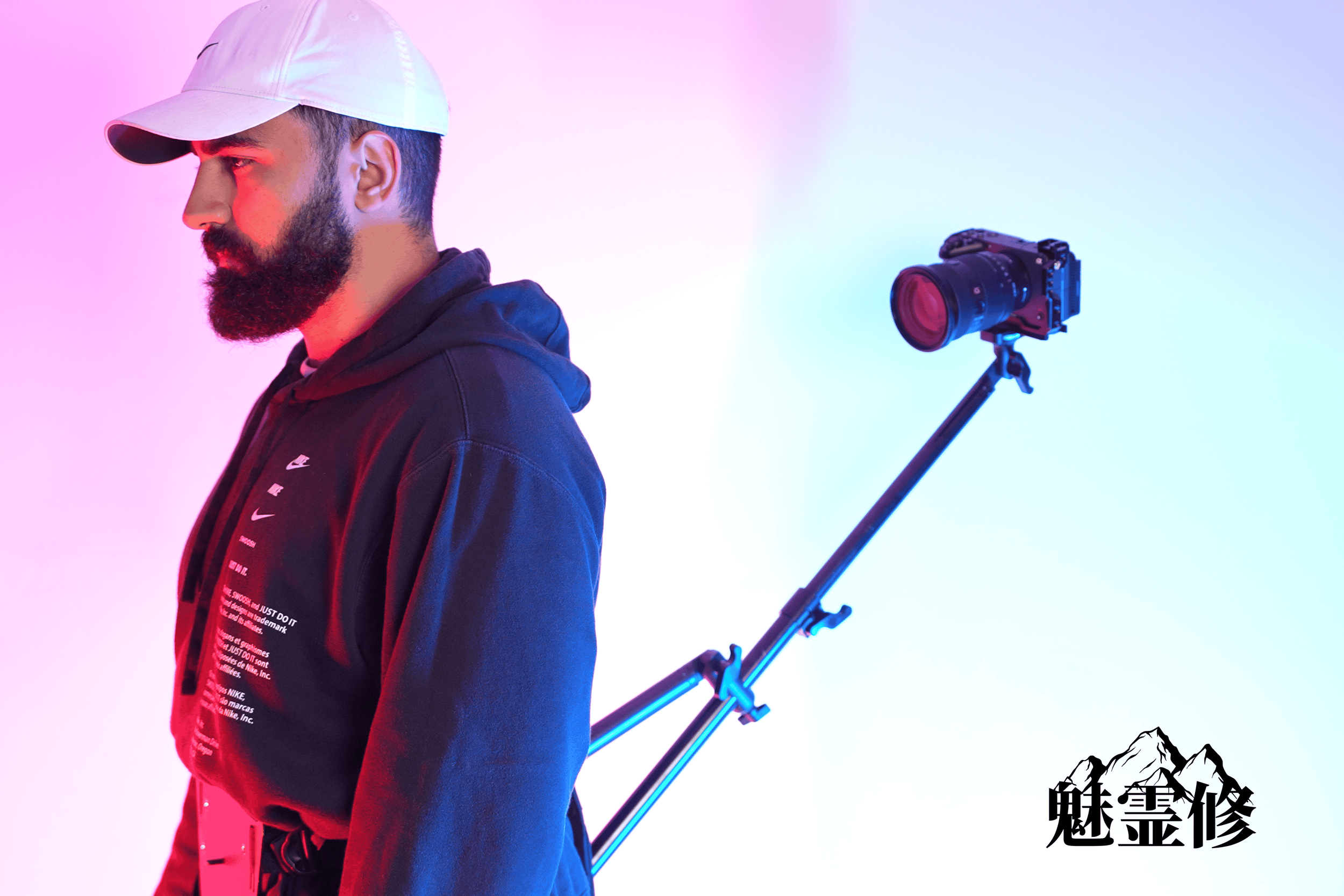 A man with a beard wearing a white cap and a black hoodie stands against a pink and blue gradient background. A professional camera on a tripod is positioned behind him, pointing towards the right. There are Chinese characters and mountain graphics i