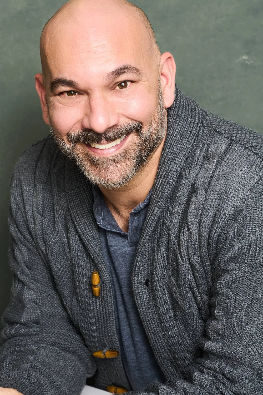A smiling middle-aged man with a bald head, beard, and mustache, wearing a gray blazer over a dark shirt against a green background.