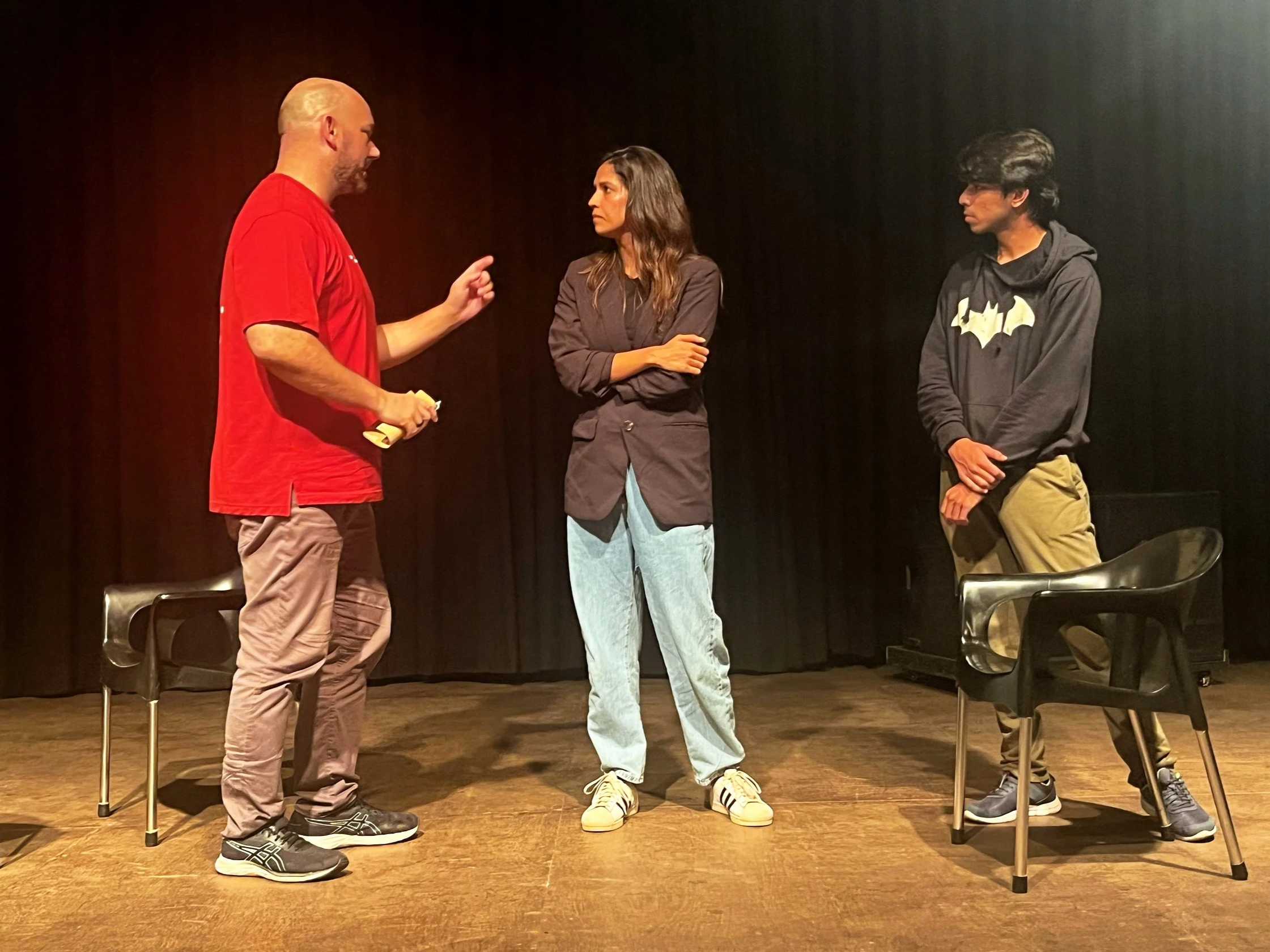 Three people stand on a stage in front of black curtains. Two men and one woman are engaged in conversation. The man on the left, wearing a red shirt and beige pants, is gesturing with his right hand. The woman in the middle, with long brown hair, wearing a dark blazer and light blue jeans, has her arms crossed. The man on the right, with curly black hair, wearing a black hoodie with a yellow bat symbol and tan pants, stands with his hands in front of him. There are two black chairs on stage, one on either side.