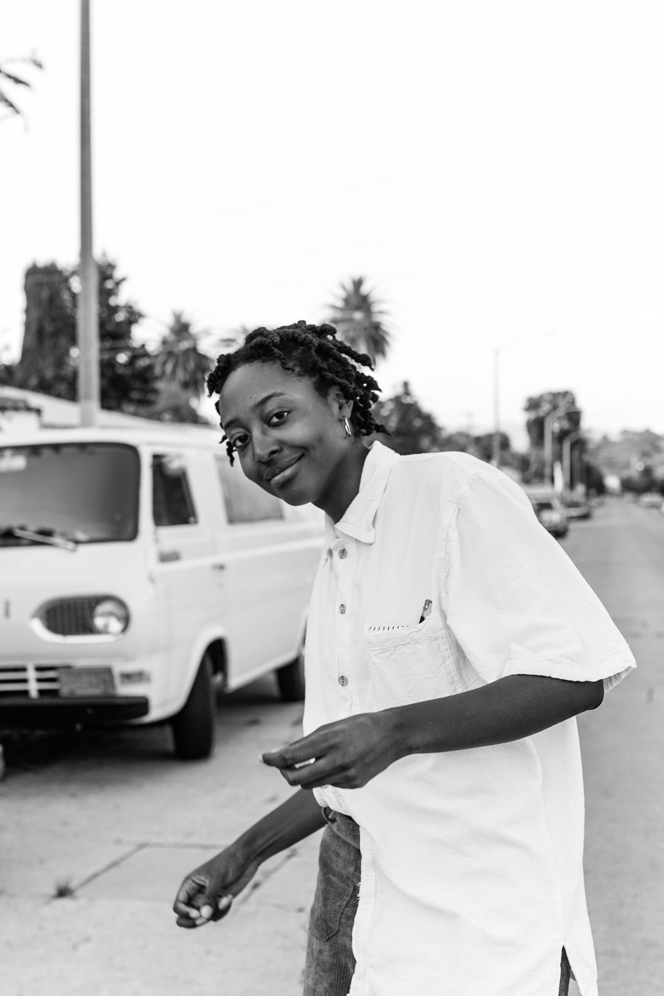 Jadonna in a white shirt standing on a street near a vintage van, with trees in the background.