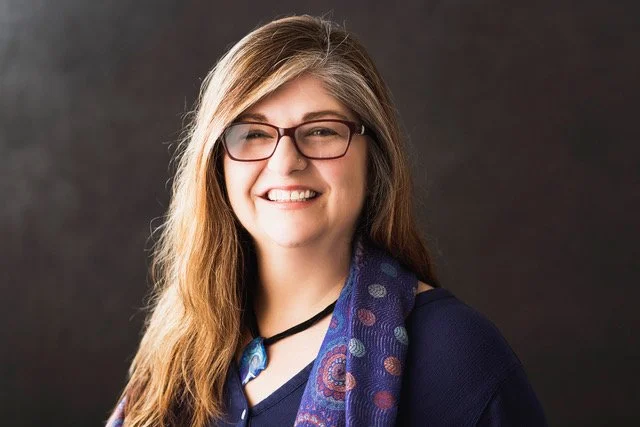 A woman with long brown hair wearing glasses and a dark blue top, smiling at the camera with a patterned scarf around her neck, against a dark background.