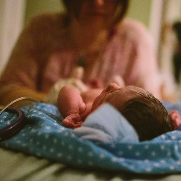 A newborn baby lying on a blue blanket with white polka dots, held by an adult woman, possibly a mother, whose face is partially visible in the background.