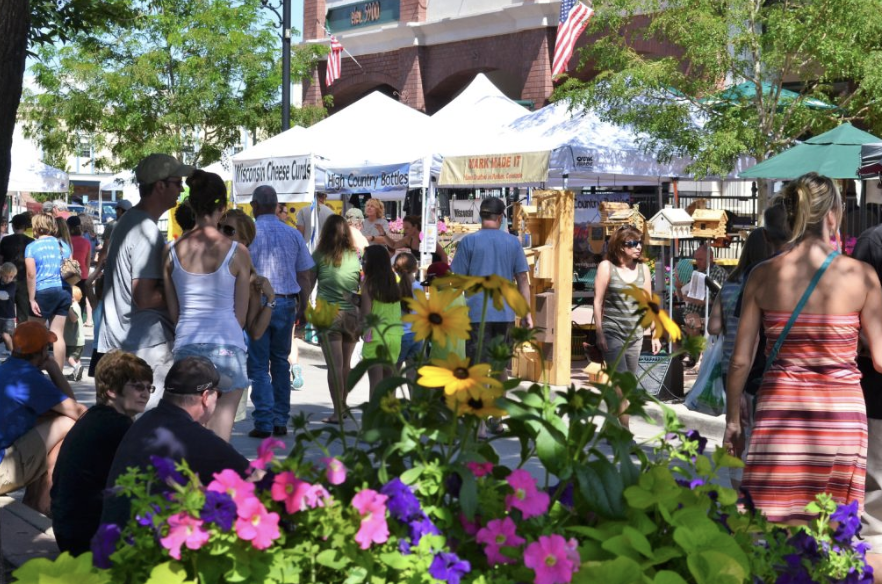 Crowd at an outdoor market with vendor tents, flowers in the foreground, and people browsing stalls under a blue sky.